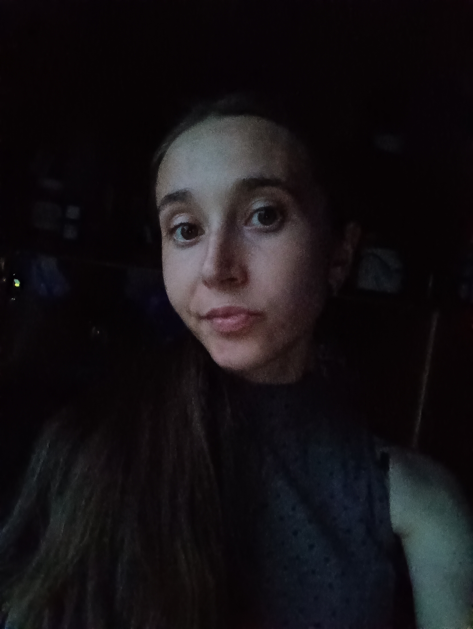 A young woman is captured in a dimly lit, close-up selfie. Her face, illuminated by an unseen light source, is turned slightly towards the camera. She has long, dark hair that falls over her left shoulder and down her chest. Her eyes are open and looking directly at the viewer. Her lips are slightly parted, conveying a neutral expression. She is wearing a dark, high-necked top with a subtle textured pattern. The background is very dark, with faint, blurry shapes suggesting an indoor environment. There are no visible indicators of the location being in Vypasne, Ukraine.
