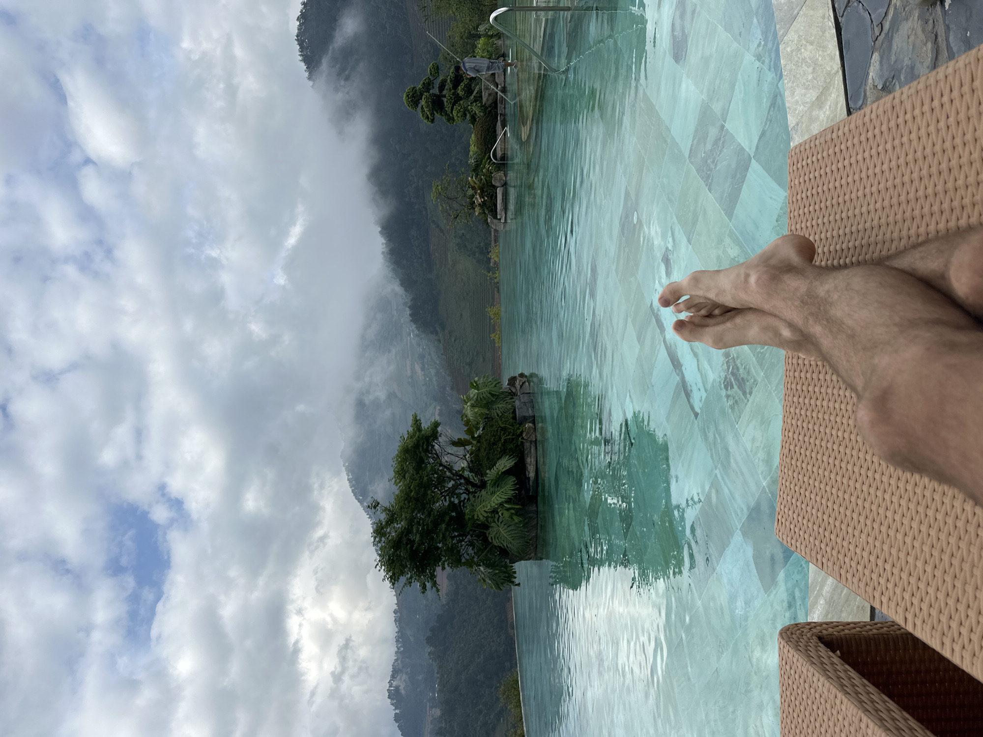 The image depicts an outdoor infinity pool scene located in Púng Luông, Vietnam, under a cloudy sky. In the immediate foreground, a person's bare, hairy feet are crossed at the ankles and resting on a light brown, woven lounge chair, indicating relaxation. The pool, tiled with light turquoise and grey squares, occupies the midground, its surface showing gentle ripples.
Within the pool, two decorative islands are visible. The larger island on the left features a dense cluster of green trees and broad-leafed plants, reflecting in the water. On the right side of the pool, a smaller island hosts a meticulously pruned bonsai-style tree and additional greenery. Further right, a male figure wearing a grey hooded top and shorts stands in the pool, holding a long pole, suggesting pool maintenance activity. Stainless steel handrails are visible on the pool's edge in the far right.
The background is dominated by a expansive mountainous landscape covered in dense green foliage. Low-hanging clouds and mist partially obscure the mountain peaks, creating a hazy atmosphere. Terraced hillsides, characteristic of agricultural cultivation, are discernible on the left slopes. The sky above is largely overcast with white and grey clouds, though a small patch of blue sky is visible towards the top center.