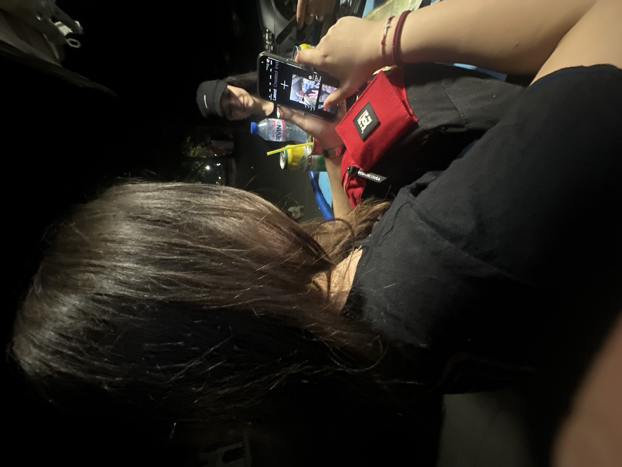 This dimly lit image captures a candid moment, likely at night in Lom, Bulgaria. The foreground shows the back of a person's head with long, dark hair. In the mid-ground, another individual's hand holds a smartphone, its screen illuminating the scene. This person wears a red bracelet and has a red pouch or wallet, featuring a white "DC Shoes" logo and the text "DC SHOECOUSA", attached nearby.
On the smartphone screen, part of an interface with "Latest Created Invent" text is visible, along with the reflected face of a third person wearing a dark cap, suggesting a video call, recording, or the camera app being active. Below the phone, a clear plastic bottle of "DEVIN" brand water and at least one silver canned drink, partially labeled "RICKO", with a yellow straw, are visible. The scene appears to be set in a casual environment, possibly inside a vehicle or a dimly lit room, with sparse background lights suggesting an outdoor context beyond the immediate subjects. The overall activity revolves around the use of the smartphone in a relaxed social setting.