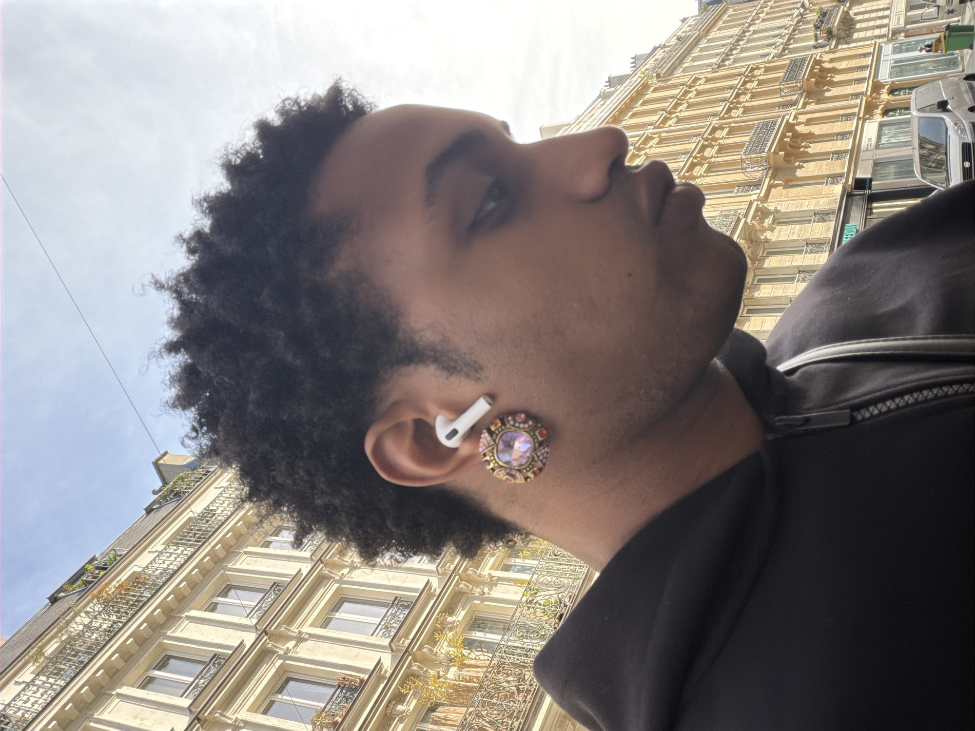 A close-up, worm's-eye view photograph captures a young person, seen from the right profile, looking upwards and slightly to the right. The individual has dark, curly hair and light facial hair stubble. A white earbud is positioned in their right ear, and below it, a large, ornate circular earring adorns the earlobe. The earring features a prominent pink or purple central gemstone surrounded by smaller, colorful gems in shades of pink, purple, orange, and gold, set within a metallic frame. The person is dressed in a dark-colored jacket or hoodie, with a zipper detail visible near the right shoulder.
The setting is an urban environment, specifically Paris, France, given the architectural style. The perspective looks up towards tall, classic Haussmannian buildings with light-colored (cream or pale yellow) facades. These buildings feature multiple stories, ornate wrought-iron balconies, and large windows. The sky above is bright and mostly clear with some faint clouds, indicating fair weather during the daytime, likely midday or early afternoon. A silver car is partially visible on the far right, situated on the street below the buildings. Faint, partial text, possibly part of a business name, is discernible on a sign on one of the buildings but is illegible due to the angle and resolution. The image conveys a casual moment of observation within the city.