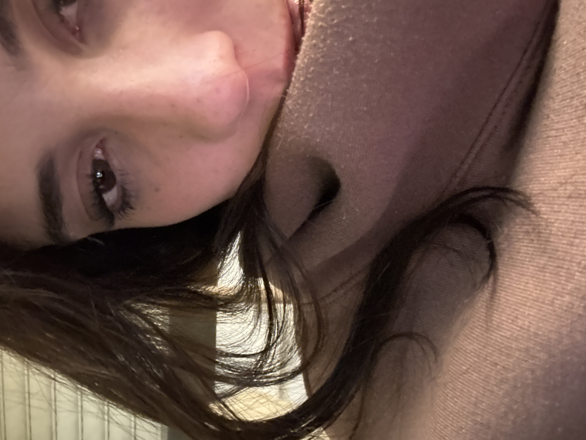 A close-up, slightly angled view features a person's face, predominantly the upper portion. The face shows details like the nose, eyes, and eyebrows, with some freckles visible. The person's hair is dark and flows around the face. They are wearing a light-colored, textured garment, possibly a sweater or hoodie. The background behind the hair appears to be a bright, neutral-colored wall with vertical linear details. The location cannot be determined from the image.