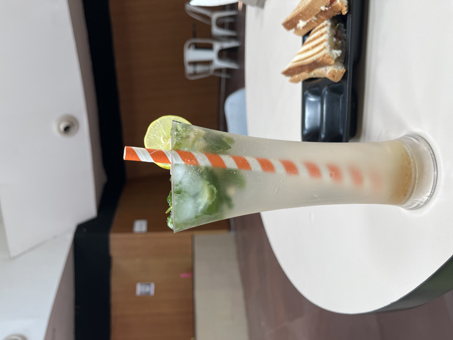 A tall glass of a refreshing beverage, possibly a mojito, sits on a white table in a modern, well-lit eatery in Sahibzada Ajit Singh Nagar, India, located at the coordinates 30.703, 76.668. The beverage is topped with a slice of lemon and a sprig of mint. A red-and-white striped straw extends into the drink. In the background, two chairs stand to the right, and behind them are more chairs and tables in a similar modern setting. Two sandwiches on black trays can be seen to the right of the glass. The scene conveys a relaxing atmosphere of a casual eatery.