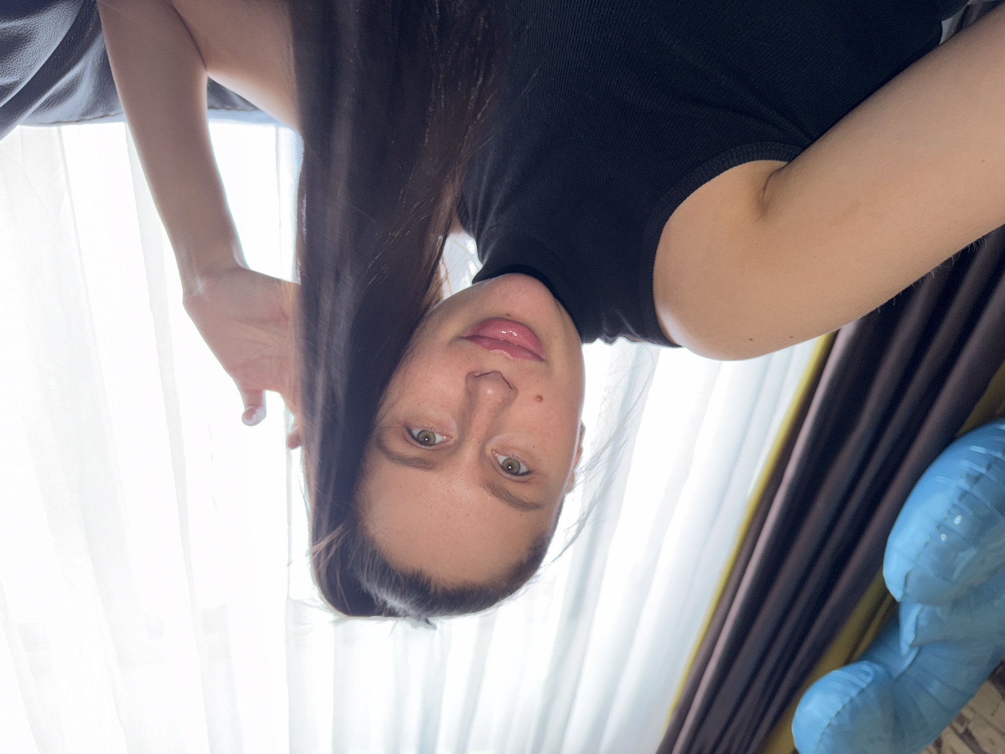 An inverted selfie captures a young woman looking directly at the camera. She has long, dark brown hair that falls over her shoulders and part of her face, fair skin, and light-colored eyes, appearing green or hazel. Her lips are slightly parted, revealing a glossy finish, and she has a subtle, gentle expression. She is wearing a black, ribbed, high-neck or turtleneck top.
Her left arm is visible, bent at the elbow, with her hand slightly curled and fingers extended towards the upper left of the frame. The background is dominated by bright natural light streaming through sheer white curtains, suggesting a window. Behind these, darker, possibly brown or gold-toned curtains are partially visible on the right. In the upper left corner, a dark, textured surface, possibly part of a sofa or headboard, is seen. To the right of the woman, a blue inflatable object, shaped somewhat like a balloon figure, is partially visible.
The lighting indicates it is daytime. The image appears to be taken indoors, likely in a room such as a living room or bedroom, with the subject holding the camera above her, resulting in the upside-down perspective. No visible text is present. While the location context is Khimki, Russia, there are no specific visual cues in the image to confirm this detail.