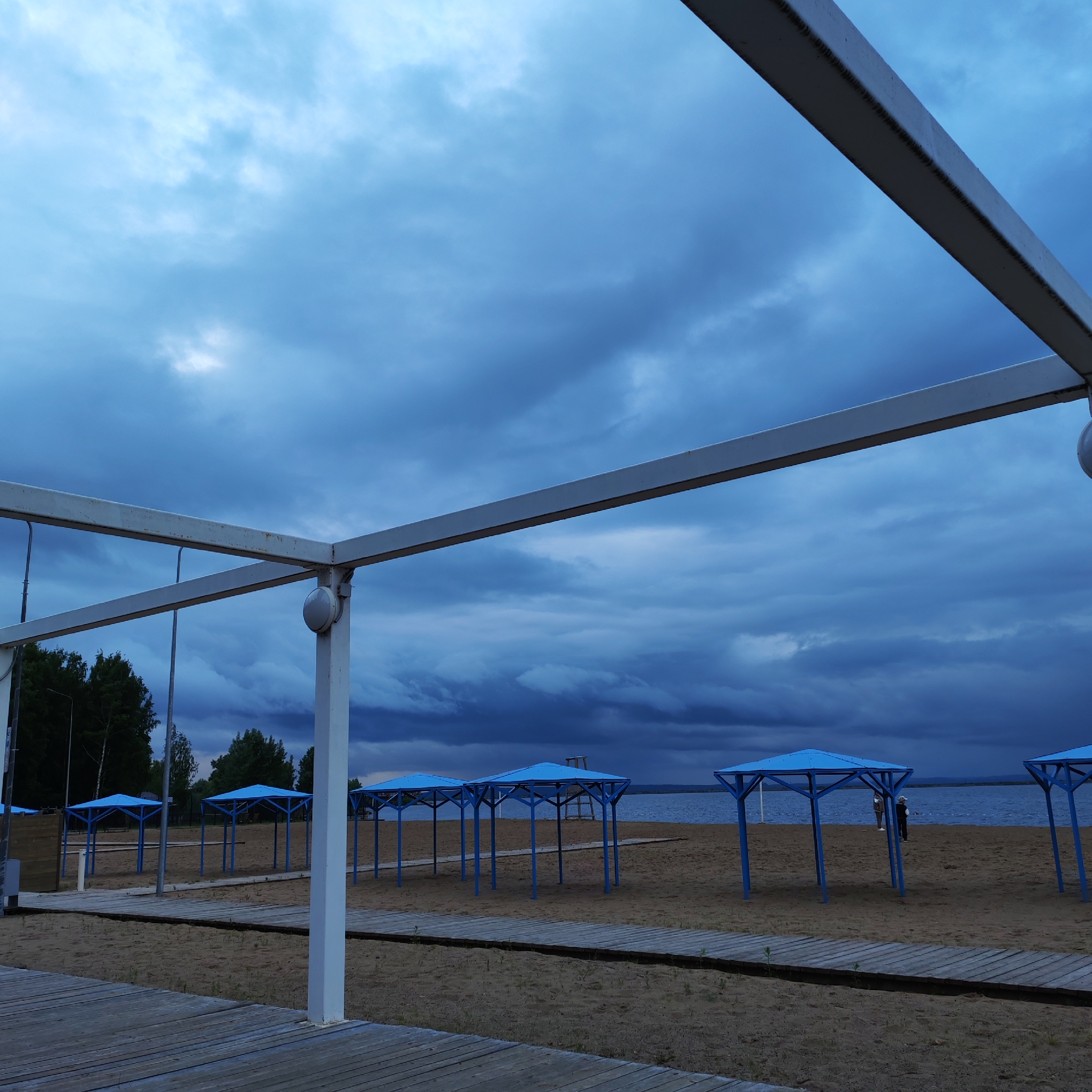 This image depicts a beach scene under heavy, dark blue-grey overcast skies, located in the city of Chaikovsky, Russia. In the immediate foreground, a weathered wooden plank walkway extends from the bottom left, partially obstructed by a white, rectangular metal frame structure. This structure consists of horizontal beams and a vertical post on the right, displaying visible weld marks and minor rust. A spherical white light fixture is affixed to the vertical post.
The midground features a sandy beach, traversed by a second, similar wooden walkway running parallel to the first, further back. Positioned on the sand are at least seven identically designed open-sided beach shelters, each with a blue rectangular roof supported by blue metal posts. A light pole is visible on the left side of the beach. Behind the shelters on the left, a dense line of dark green deciduous trees forms the tree line.
In the background, a wide body of dark blue-grey water, consistent with a large lake or reservoir, stretches across the horizon. Distant land masses are faintly visible on the far right across the water. On the beach near the water's edge, between the shelters, two indistinct human figures are visible; one stands facing the water, while the other, closer to a blue shelter, appears to be standing or slightly bent. A tall, white lifeguard tower is discernible in the distance on the beach to the right of the leftmost blue shelter. The sky above is completely covered by dense, textured, dark blue and grey clouds, with some lighter patches suggesting diffused light penetration. The overall lighting is dim, indicating heavy overcast conditions or dusk.