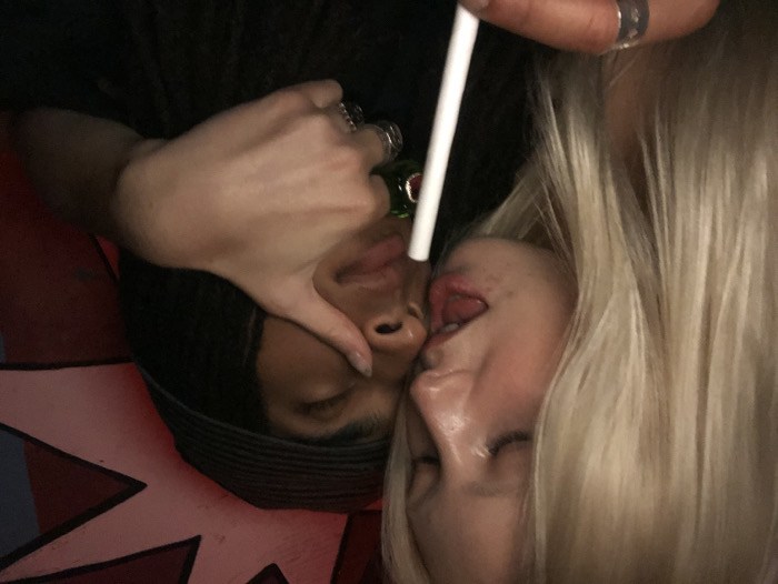 An intimate, close-up shot captures two individuals in a dimly lit setting, likely at night. The image is tilted, suggesting the subjects are lying down or captured from an unconventional angle.
On the left, a person with dark skin and short dark hair (possibly styled in waves or braids under a head covering) has their eyes closed. A hand, adorned with multiple rings, gently cups their face or chin. This hand appears to belong to the second individual.
On the right, a person with fair skin and long, flowing blonde hair is featured. Their eyes are open, and their tongue is slightly extended, giving a playful or sensual expression. There is visible moisture around their mouth and chin.
A white straw or stick is held by a hand (likely the dark-haired person's) near the blonde-haired person's face, with a small green bottle visible behind it, suggesting they might be sharing a drink. The two individuals are in very close proximity, indicating a moment of intimacy or affection.
The background is largely dark, with a striking red and white graphic pattern, resembling a stylized star or explosion, visible in the lower left corner. The overall mood is intimate and possibly flirtatious. No text is visible in the media file. The location context provided is Paris, France.