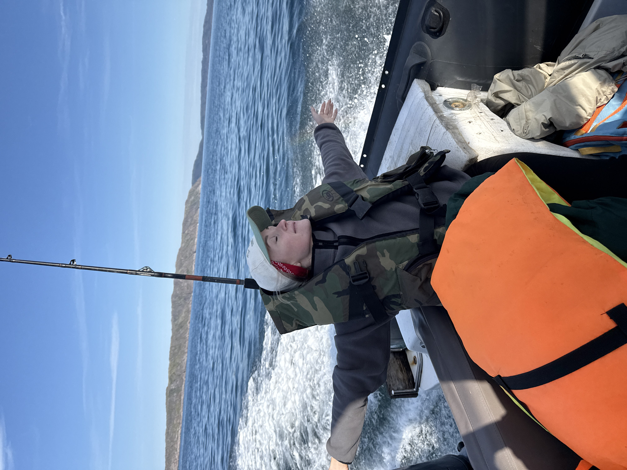 A person with eyes closed and arms outstretched is on a boat, enjoying the ride. They are wearing a life vest over a gray jacket and a baseball cap. The boat is moving, creating a wake in the blue water. In the background, there are hills and a clear sky. The scene appears to be in Teriberka, Russia.