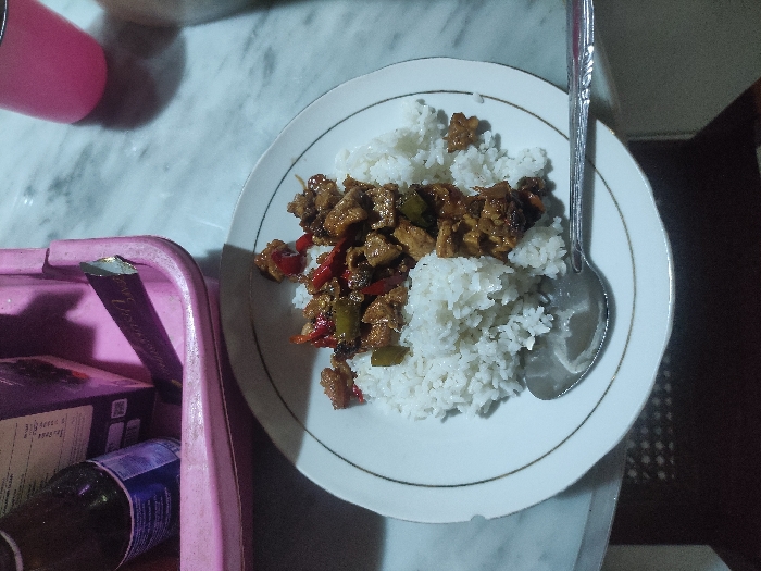 A white plate with a portion of white rice and a stir fry with meat, vegetables, and sauce. A stainless steel spoon is lying next to the plate, with the handle extending off the right side of the image. The plate sits on a white marble table with a partially visible pink plastic container in the upper left corner of the frame. The location is Mojokerto, Indonesia (latitude: -7.459, longitude: 112.447).