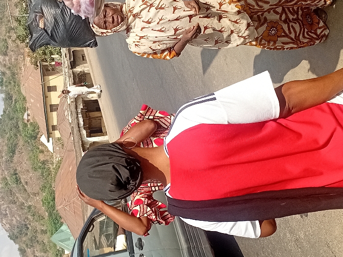 A woman carries a bag on her head while standing next to an older woman in patterned clothing, who also carries a bag on her head. Another woman in white shorts and a red top stands with her back to the camera. They appear to be on a roadside with buildings and a hillside in the background. The photo is taken at an angle.