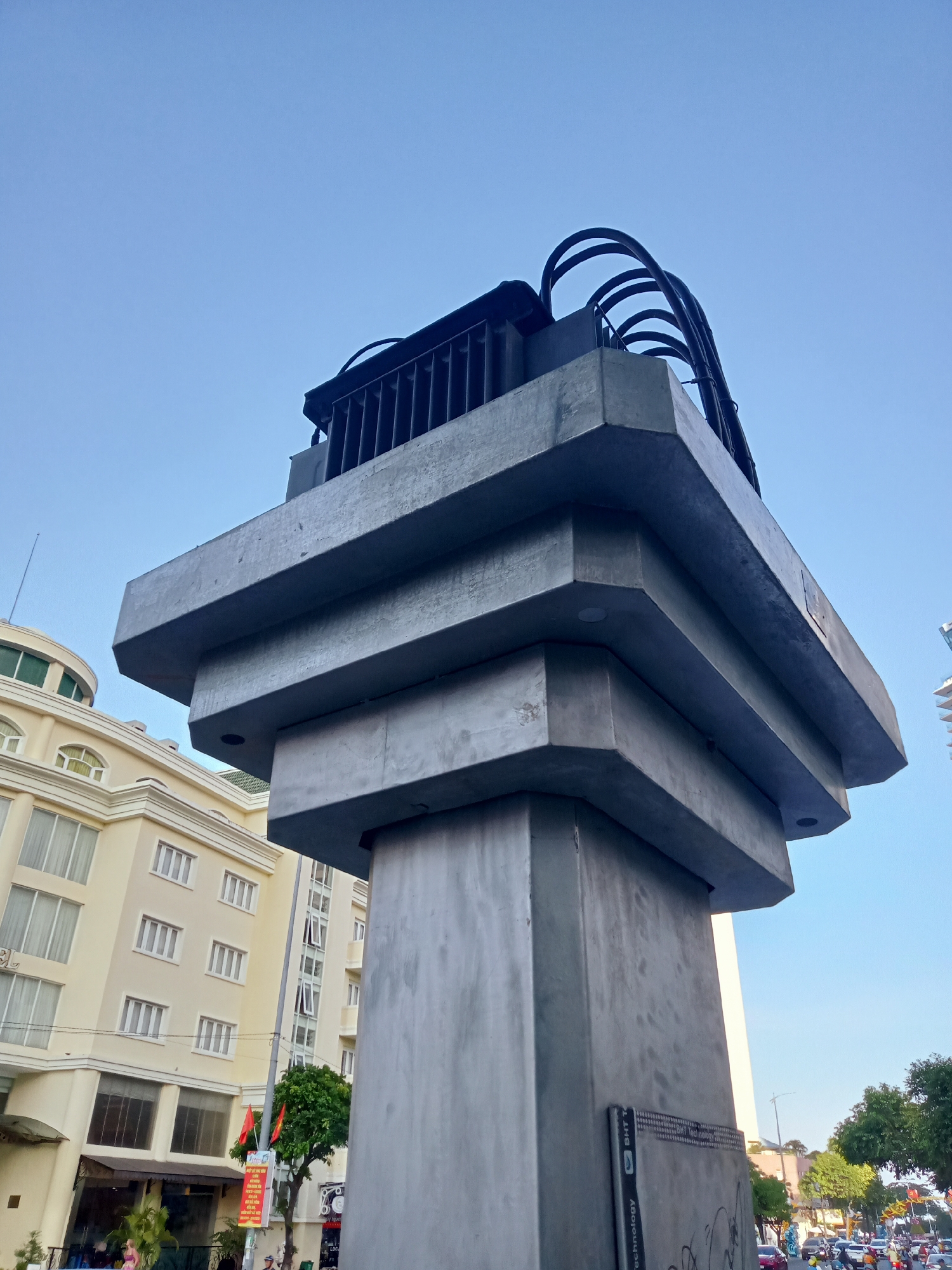 The image shows a tall, concrete structure or pillar in Nha Trang Ward, Vietnam. The pillar is composed of stacked, rectangular blocks that gradually widen towards the top. At the peak of the structure, there is a black, cage-like enclosure with curved, metal supports. The base of the pillar features graffiti and a sticker. In the background, a multi-story, tan-colored building with multiple windows is visible, along with other buildings, trees, and street traffic. The sky is clear and blue.