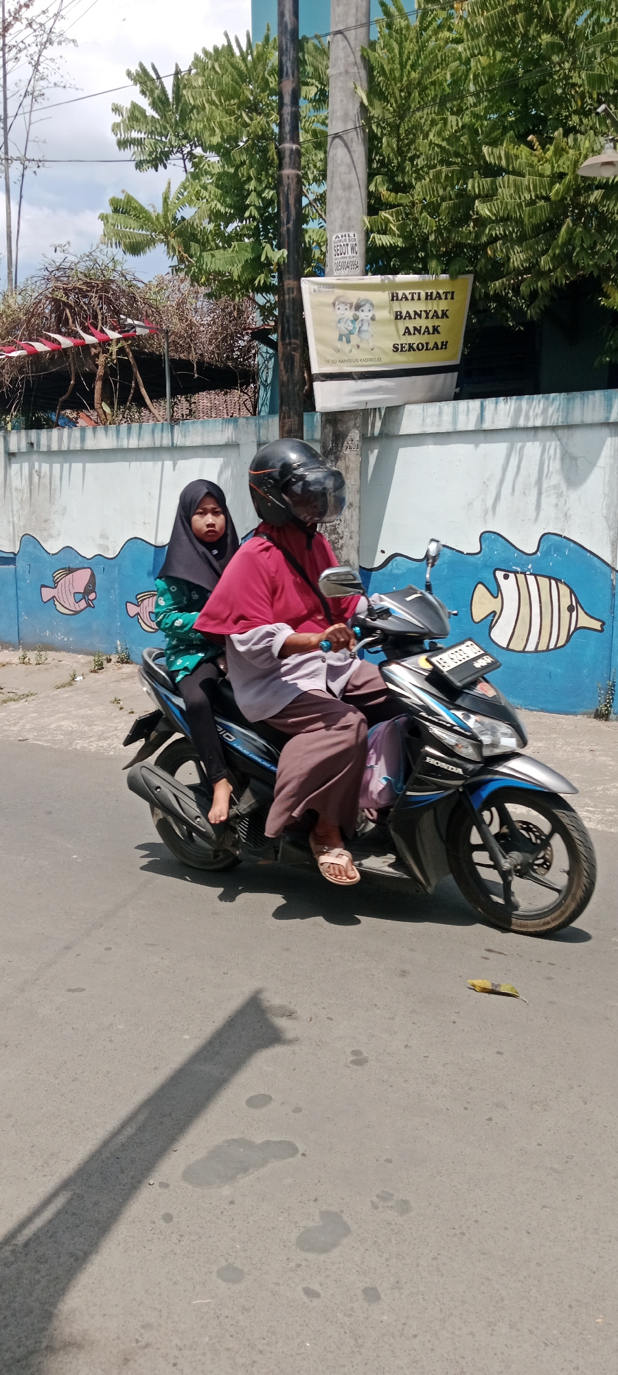Two individuals, an adult and a child, are riding a motorcycle on a paved road in Purwomartani, Indonesia. The adult, wearing a red head covering and a light brown long-sleeved shirt with a maroon outer garment, is driving the motorcycle. They have a black helmet with a clear visor. The child, wearing a black hijab and a green patterned shirt, is seated behind the adult. The motorcycle is dark colored with blue accents and has a license plate visible. In the background, there is a concrete wall with a mural of stylized fish. Behind the wall, trees and foliage are visible. A utility pole stands to the left of the wall. A sign with text and illustrations of children is attached to the utility pole. The ground is asphalt and there are shadows cast on it. A single yellow leaf is on the road in the lower right quadrant of the image.