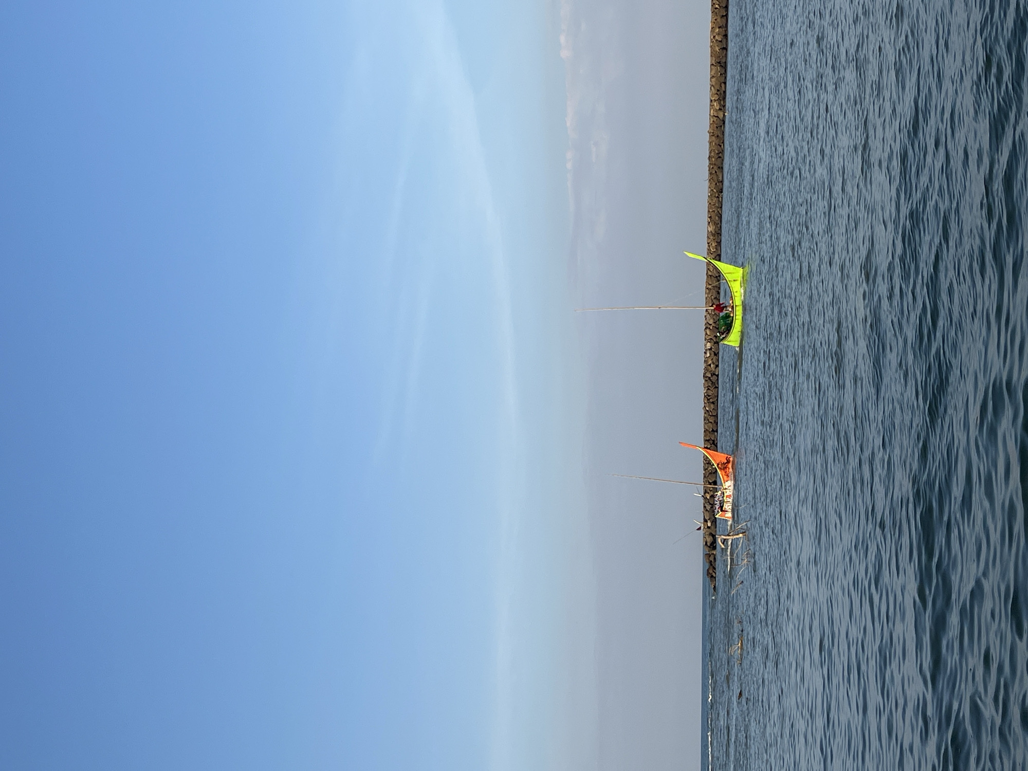Two distinct, brightly colored boats are positioned on a body of water with a clear blue sky overhead. The boat on the left is predominantly orange with a tall, slender mast and appears to be partially submerged or listing, with visible debris around it. The boat on the right is lime green, also with a mast, and has at least two individuals onboard. A long, dark, stony structure, likely a breakwater or jetty, runs horizontally across the middle ground of the image, separating the boats from a calmer area of water. The water itself is a deep blue, rippling with moderate waves, suggesting an open body of water such as a sea or large lake. The horizon is visible as a faint line between the water and the sky, with a subtle haziness. The overall scene suggests a recreational or working maritime environment, possibly near a coastline. The location is identified as Reudeuep, Indonesia.