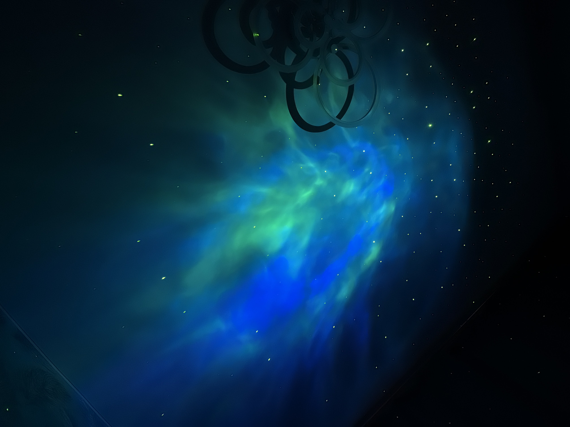 The image presents a dark, abstract scene illuminated by blue and green nebulous light. A series of dark rings are present in the upper portion of the image, possibly a decorative structure. Small, bright, yellow-green dots, resembling stars or small lights, are scattered throughout the dark areas. The origin of the image is unknown. There is no information available about the city of Reshetnikovo, Russia in this image.