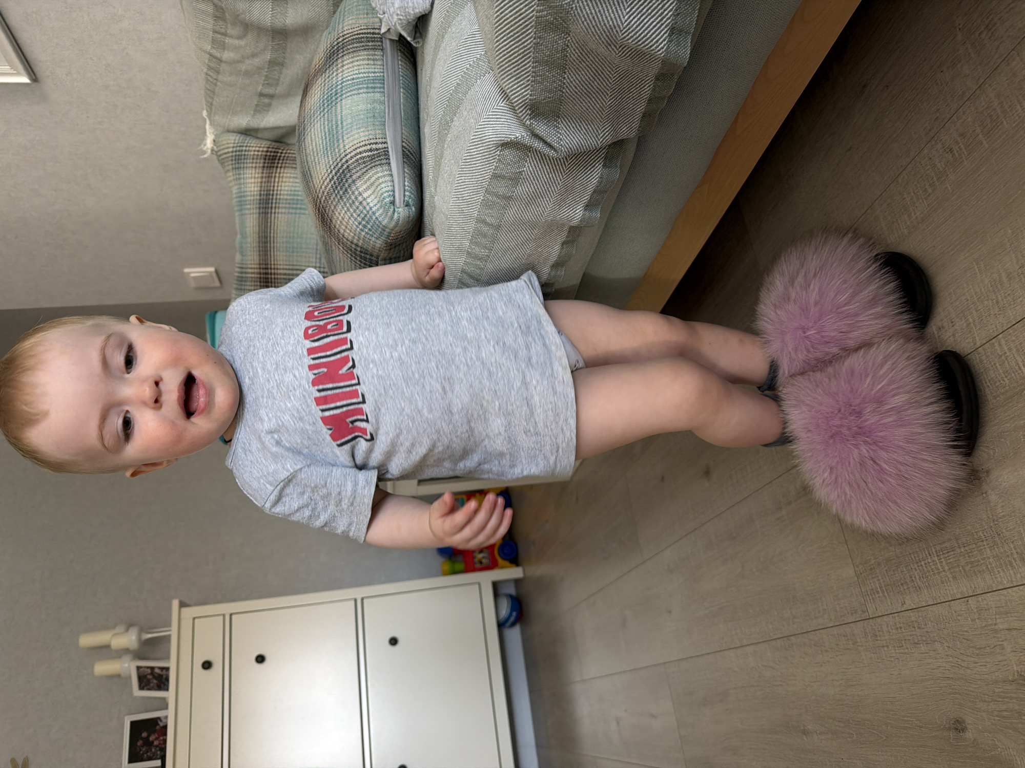 A small child with blonde hair, wearing a gray t-shirt, stands on a wooden floor with light pink furry slippers. The child is facing the camera and appears to be talking. Behind the child is a white cabinet and a portion of a light-colored couch with pillows. A toy is partially visible on the cabinet. The location appears to be indoors, likely a home. The city and country are Votkinsk, Russia.