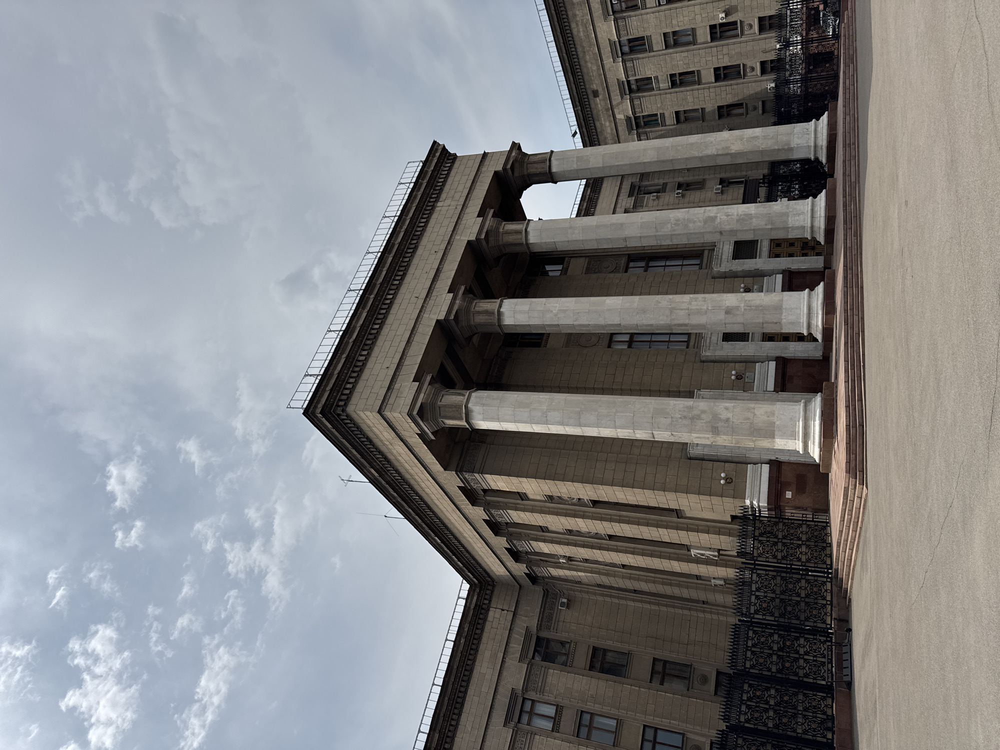 A wide-angle, low-angle exterior shot captures a monumental, neoclassical building under a partly cloudy sky. The building is constructed of light brown stone blocks and features a prominent portico with four massive, fluted columns. The columns support a substantial entablature and a cantilevered roof. The building's facade is symmetrical, with evenly spaced windows with dark frames on multiple levels. A black, ornate metal fence runs along the front of the property, separating it from a paved open area. The ground in front of the building is a light grey asphalt. The scene suggests an official or governmental institution, likely in a city center.
The building is the Central State Museum of Kazakhstan in Almaty.