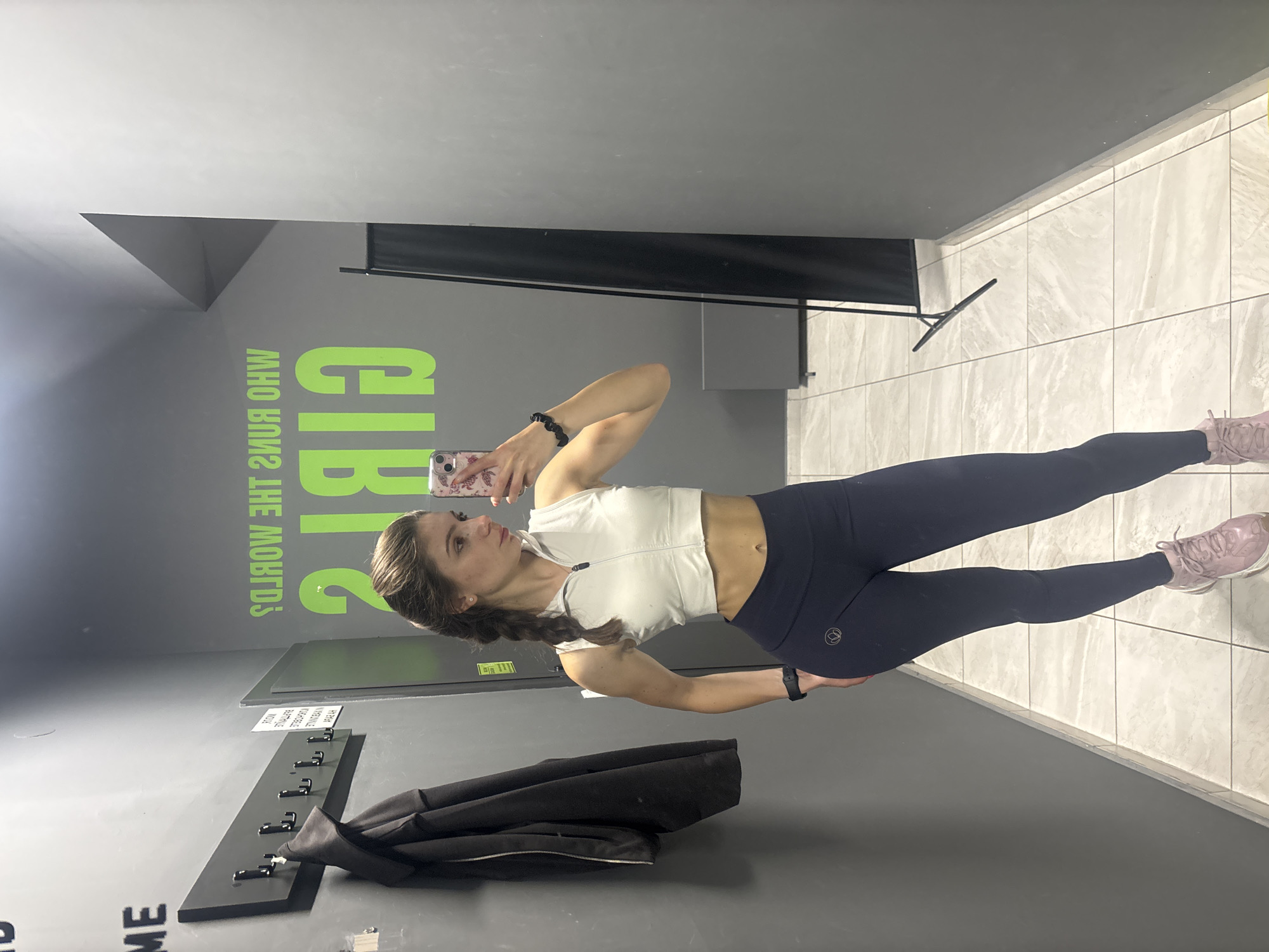 A woman stands in a narrow space with grey walls and light-colored tile flooring. She is facing a wall with a mirror and is taking a selfie with a smartphone. She is wearing a white athletic top and dark leggings. She has her hair in a braid. A black jacket hangs on hooks on the wall. Green lettering reading "Girls, Who Runs The World?" is visible on the wall. There are no other people in the image. The location appears to be a changing room in a gym. Specific location in Varna is not identifiable from this image.