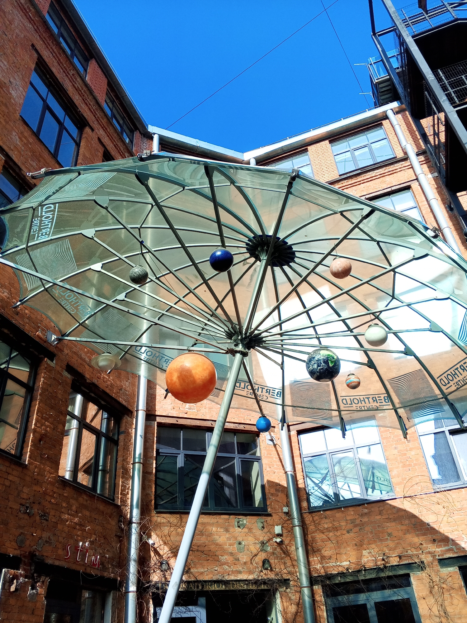 The image displays an urban courtyard or creative space within the Berthold Centre, located at Grazhdanskaya Ulitsa, 13-15, Saint Petersburg, Russia. The scene is observed from a low-angle perspective, looking upwards toward the sky.
A large, inverted, umbrella-like structure with a transparent, corrugated canopy dominates the central view. This canopy, supported by a metallic framework and a central pole, reflects the surrounding brick buildings and the reversed text "BERTHOLD CENTRE" multiple times. Suspended from the underside of the canopy are multiple spherical models of varying sizes and colors, representing planets, including blue, orange, grey, and a globe depicting Earth.
The space is enclosed by multi-story buildings constructed of reddish-brown brick. These buildings feature numerous rectangular windows with dark frames, some reflecting the canopy and the clear blue sky overhead. On the right side, an external black metal staircase or fire escape structure is affixed to one of the brick buildings. Vertical silver-colored pipes run along the brick walls. Sparse, dried climbing vines are visible on various sections of the brickwork, and faded red lettering, potentially "stim," is partially discernible on a lower wall.
The environment is brightly lit by direct sunlight, evidenced by distinct shadows cast on the brick walls and the structural elements of the canopy. The sky above is clear and blue. No individuals are present in the depicted scene.