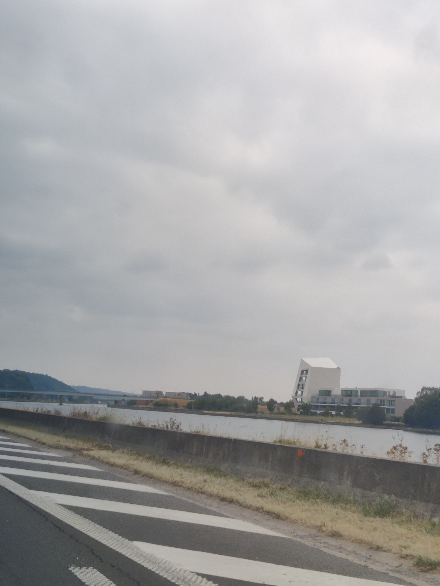 A grey, overcast sky dominates the upper portion of the image. In the distance, a body of water, likely a canal or river, is visible, with buildings and trees lining its banks. A prominent modern white building with an angular design stands out on the right side of the water. A bridge spans the water further in the background, connecting the two sides. The foreground of the image features a road with clear white lane markings and a concrete barrier. The scene appears to be a view from a road adjacent to a waterway in or near Hermalle-sous-Argenteau, Belgium.