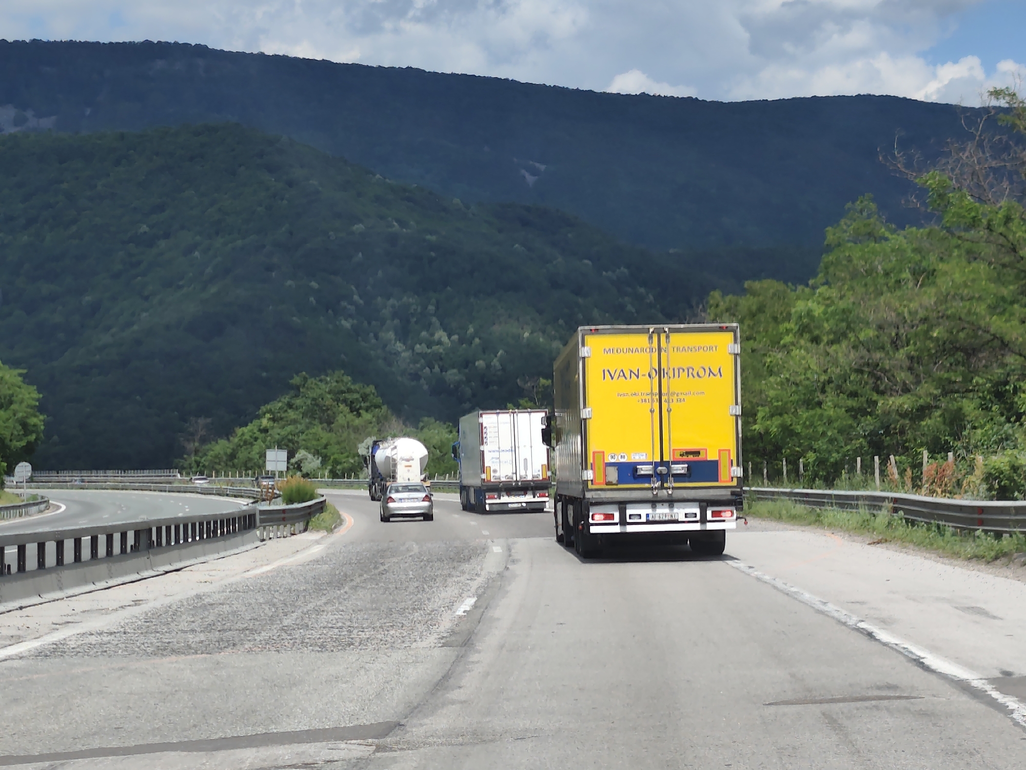 The image captures a scene on a highway near Vrachesh, Bulgaria, likely during daylight hours. The foreground shows a paved road with visible lane markings and some wear and tear. A bright yellow truck with "IVAN-OKIPROM" and "MEZHDUNARODEN TRANSPORT" written on its side is prominently featured in the right lane, moving away from the viewer. Behind it, in the same lane, is a white refrigerated trailer truck. The left lane shows a silver car driving ahead of a white tanker truck. The road curves to the left, with metal guardrails lining the median and the right side of the road. Beyond the road, a lush, tree-covered mountain range dominates the background. The sky is partly cloudy. The scene depicts traffic on a highway amidst a natural, mountainous landscape.