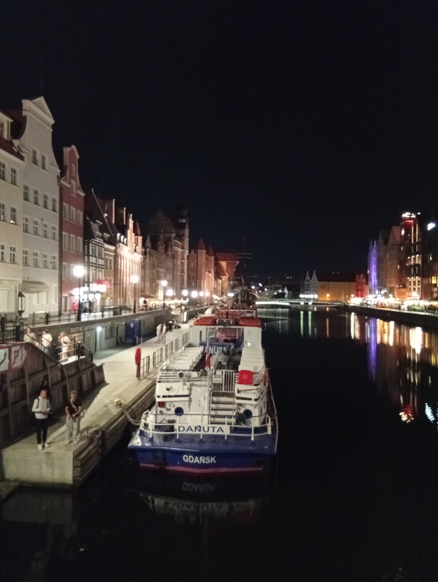 This image depicts a nighttime scene along the Motława River in Gdańsk, Poland. The riverfront is lined with historic, colorful buildings with illuminated windows, characteristic of the Old Town. A white and blue passenger boat, named "GDANSK," is docked in the foreground, reflecting in the dark water. Several people are visible on the boardwalk and near the boat. The sky is dark, indicating it is night. The overall atmosphere is one of urban tranquility and historical charm.