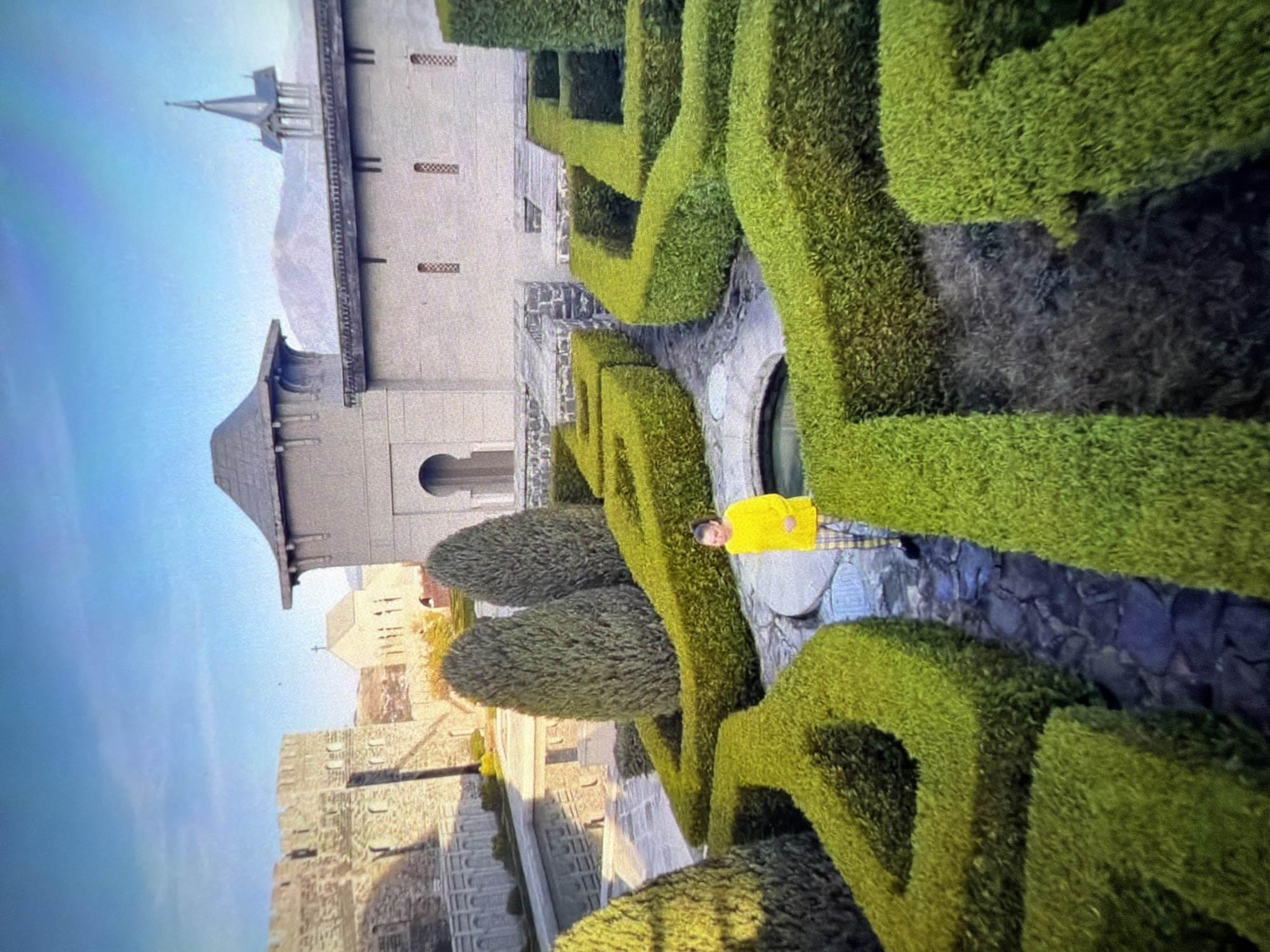 The image shows a person in a yellow shirt standing in a green hedge maze. In the background is a multi-tiered stone building with a pointed tower. The location is in the city of Unknown, Unknown.