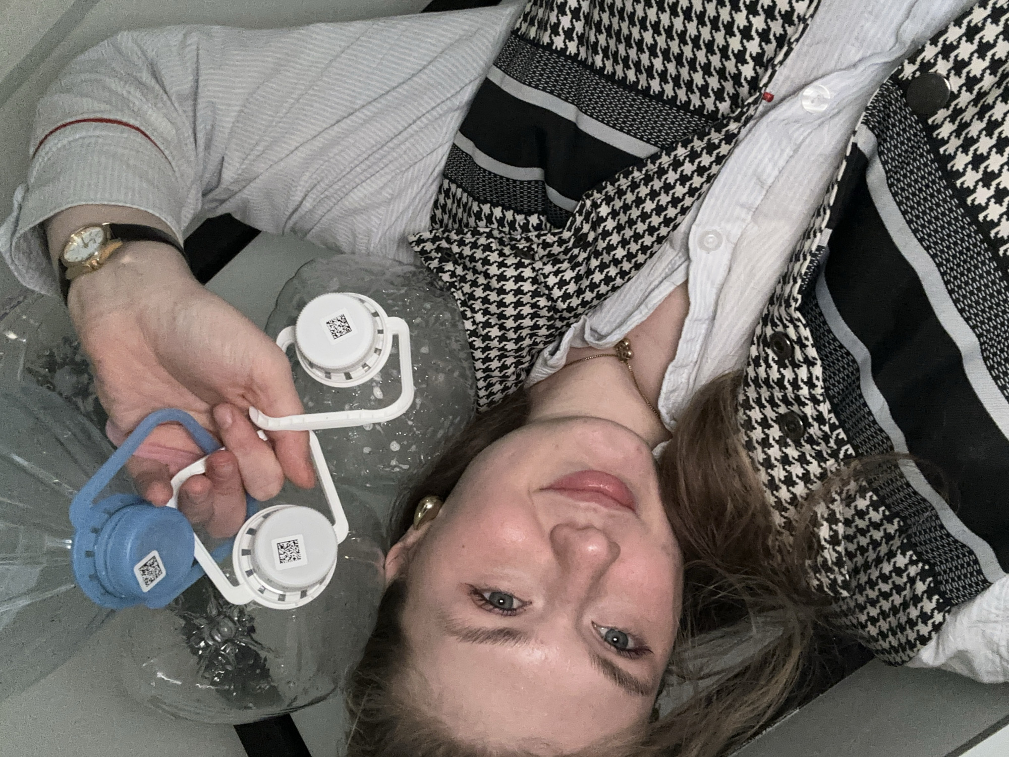 A person lies on their back with a large clear bottle containing water and two bottle caps, one white and one blue, connected by a plastic handle. The person is wearing a light-colored button-down shirt, a black-and-white houndstooth vest, and a gold wristwatch on their left wrist. The photo was taken from above, with the person looking up at the camera. I cannot place the exact location to Kazan, Russia from this image.