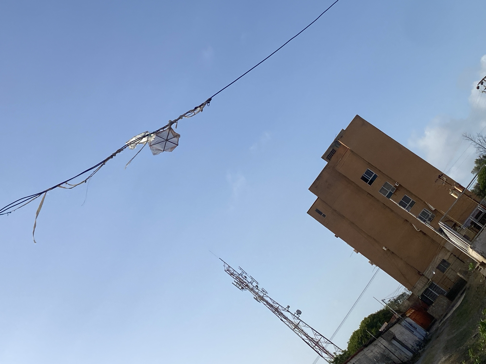 The image shows a tilted view of a cityscape in Juan Griego, Venezuela. An old, possibly damaged, geometric kite is caught on a high-tension power line that stretches across the upper part of the frame. A tall, light brown building is located on the right side of the image. A communication tower is visible below the kite and in front of the building. The sky is light blue with minimal cloud cover. A wall and some vegetation are visible at the bottom-right corner.