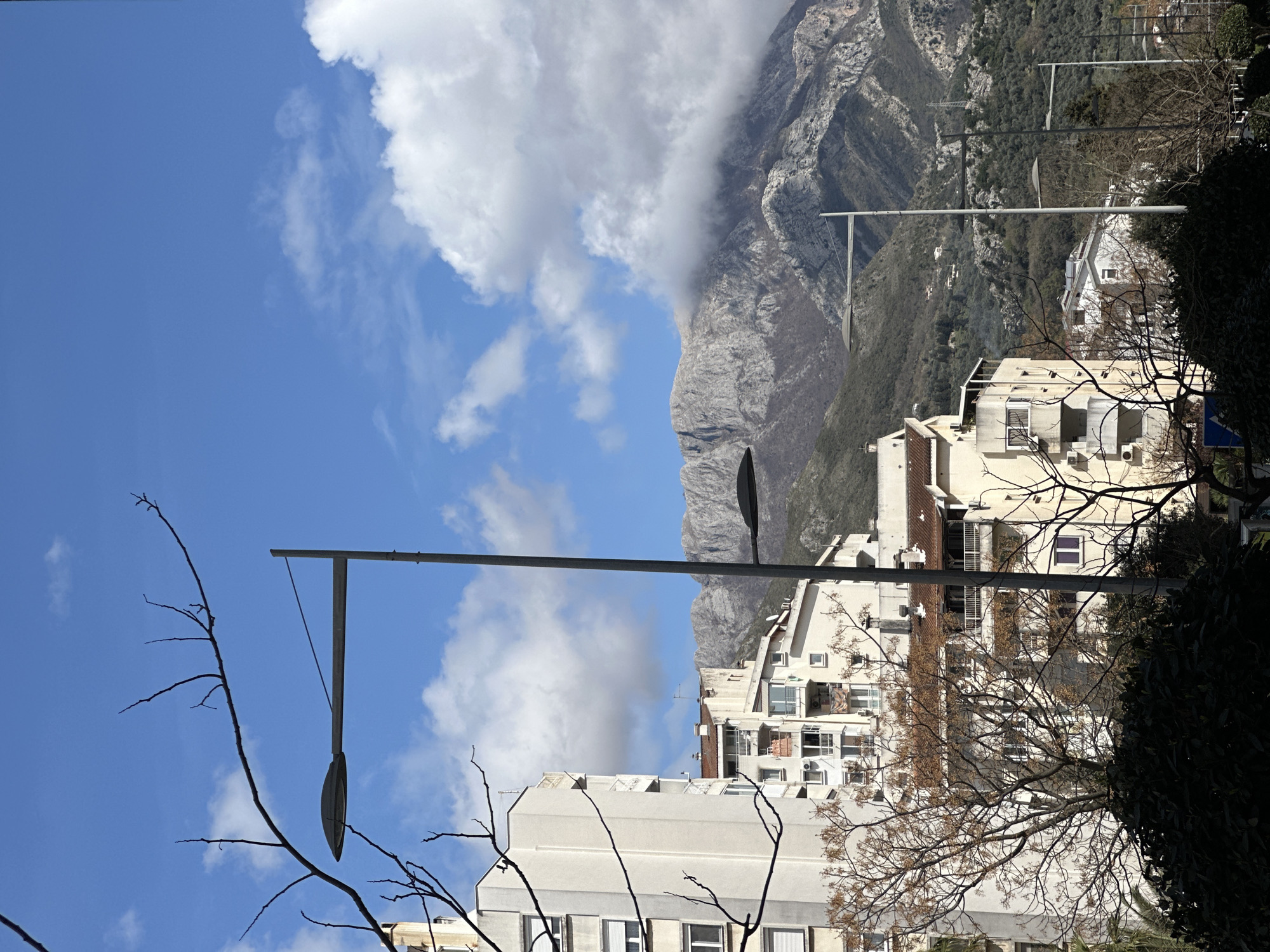 The image depicts a view in Šušanj, Montenegro, with buildings and a mountain range visible against a blue sky with clouds. A street lamp is in the foreground, partially obscured by bare tree branches. The buildings are light-colored and multi-story, with visible windows and balconies. The mountain range is rugged and partially covered by clouds.