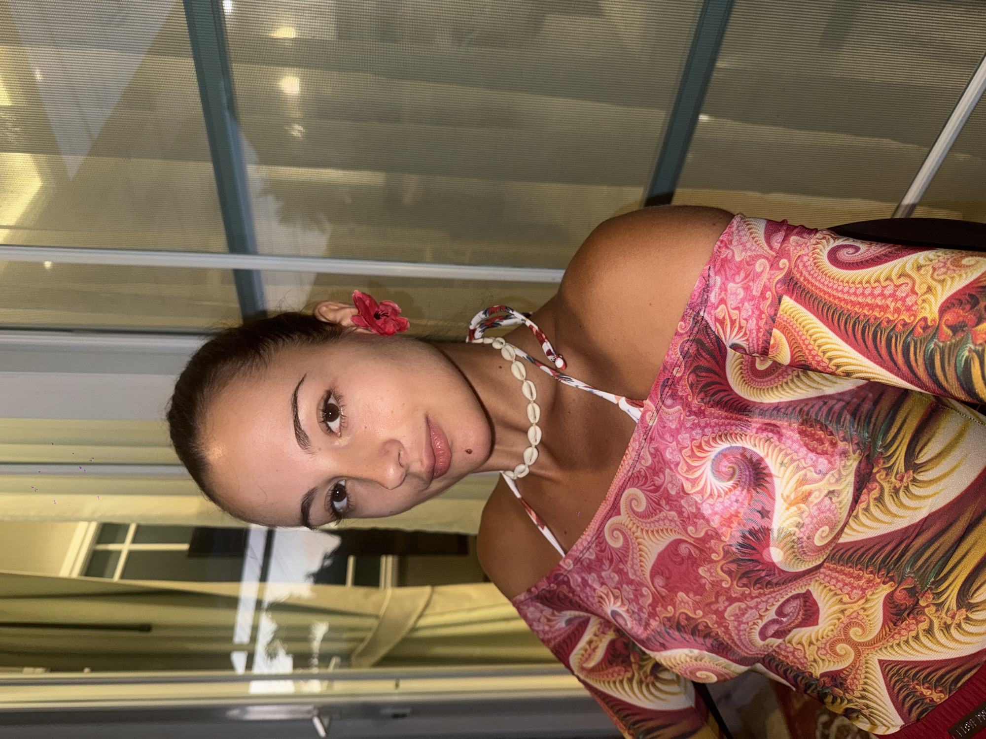 A woman is seen indoors in an apparent beach setting. She is adorned with a red flower tucked behind her ear, and she is wearing a shell necklace. Her clothing appears to be patterned and multicolored. The background suggests a structure with reflective or translucent panels. The scene is most likely not set in the city of Chalong, Thailand. The environment suggests an indoor location mimicking an outdoor, perhaps tropical, setting.