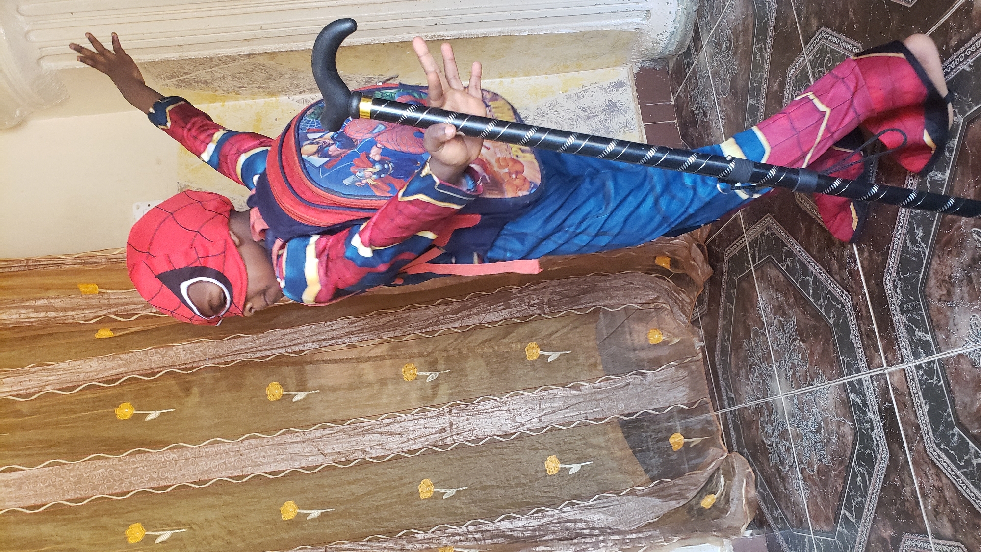 A person in a Spiderman costume stands on brown tiled flooring. They are holding a black walking stick with a curved handle. The person is positioned near a bed covered with a gold-colored fabric featuring a floral design. The scene appears to be indoors, potentially a room in a house or apartment. It is located in Narayi, Nigeria.