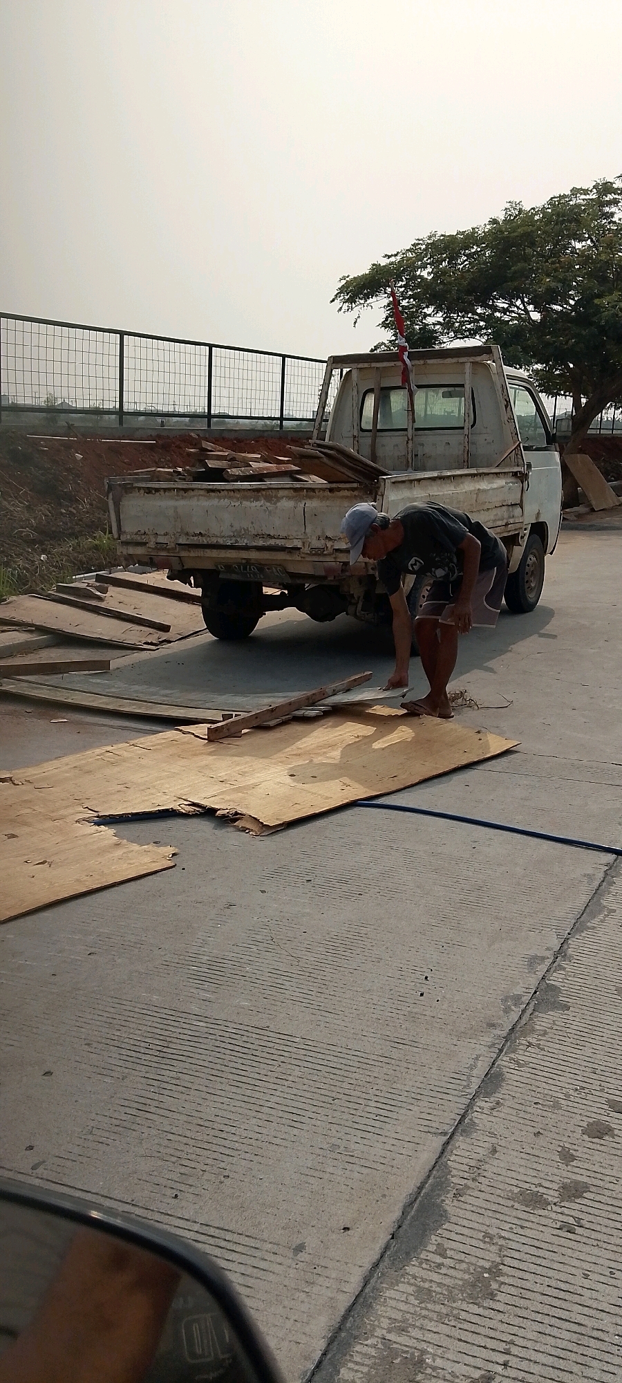 In the city of Satriamekar, Indonesia, a man is seen crouching in front of a white pickup truck laden with wooden debris. The truck, parked on a concrete surface marked with parallel lines, is partially loaded with planks of wood. The man, wearing a dark t-shirt and shorts, appears to be interacting with a large sheet of plywood on the ground in front of the truck. Behind the truck, a chain-link fence separates the immediate area from a reddish-brown dirt mound and some vegetation. A tree with dense foliage is visible to the right of the truck, and a red and white flag is mounted on its rear. The sky is overcast, and the overall lighting suggests it is daytime. In the foreground, a car's side mirror is visible, reflecting a portion of the scene.