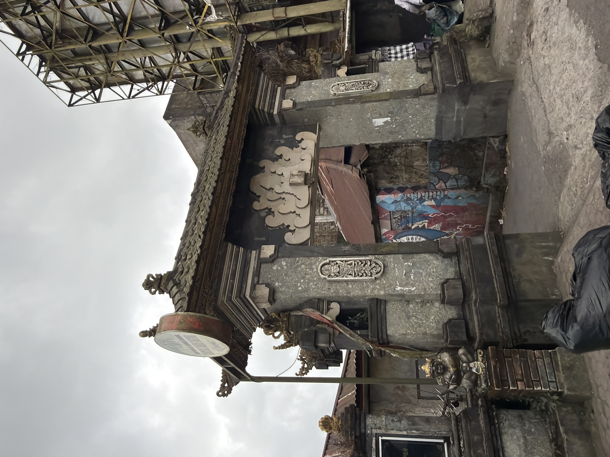 The image shows a street scene in Kintamani, Indonesia. In the foreground, a grey stone gateway with traditional Balinese architectural elements serves as the entrance to a building or complex. The gateway is adorned with decorative carvings and topped with a tiled roof. A circular sign hangs from the left side of the gateway. The interior of the gateway features graffiti art and a makeshift roof made of corrugated metal. To the right of the gateway, a large metal billboard structure is visible. The ground is paved with stone and dirt, and several black trash bags are present in the lower portion of the image. The sky is overcast.