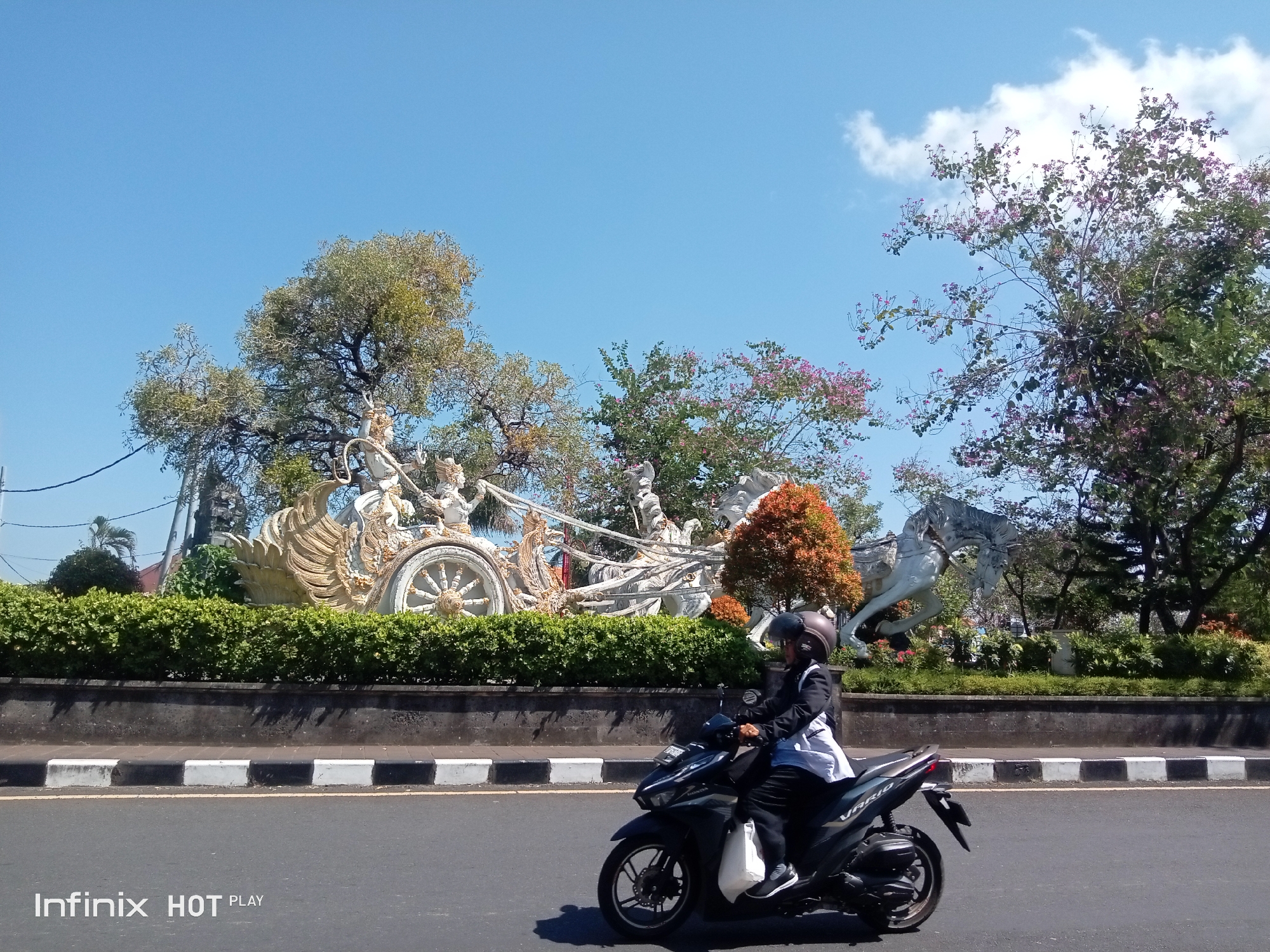 A person rides a dark-colored motorcycle with a white bag on the right side, in the foreground, along a paved road with a white and black striped curb. The rider wears a helmet and a black and white outfit. In the background, a large, ornate white sculpture depicting a chariot with horses and human figures is visible, set against a backdrop of lush green bushes and trees with pink flowers. The sky is clear and blue with scattered white clouds. The scene appears to be in Gianyar, Indonesia, indicated by the "Infinix HOT PLAY" watermark in the bottom left corner, a common branding on devices sold in the region.