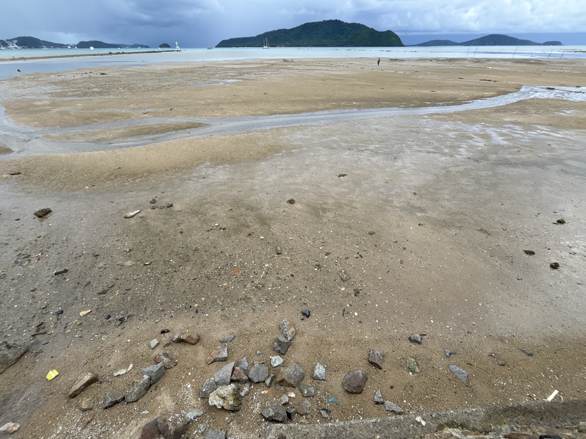 A wide, low-angle, outdoor, daylight, panoramic shot shows a tidal beach area in Rawai, Thailand, during low tide. The foreground is dominated by wet sand, small stones, and scattered debris, including a yellow plastic wrapper and blue plastic fragments. In the midground, the exposed sand exhibits patterns of receding water, creating shallow pools and rivulets. Further in the distance, several small sailboats are visible on the water, and a single person is walking on the beach. The background features a verdant, tree-covered island with a lighthouse on its far side, under a cloudy, overcast sky. The overall scene suggests a humid, tropical environment.