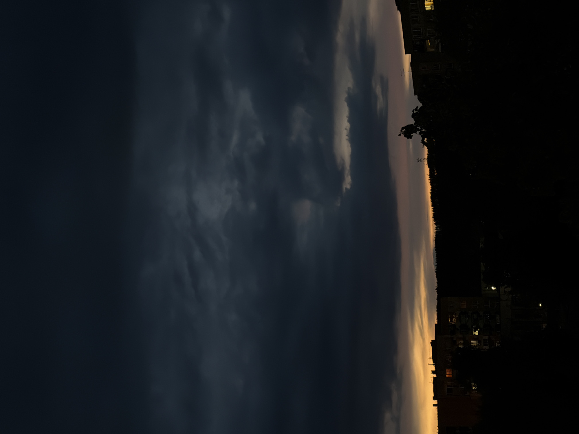 The image depicts a darkening evening scene in an unidentified location within an unknown city. Dominating the left side of the frame is a heavy, dark cloud cover, transitioning into lighter, illuminated clouds towards the center. Along the horizon, a thin band of golden light remains from the setting sun. To the right of the illuminated sky, there's a dark silhouette of trees and buildings. Some buildings have lights on, suggesting the onset of night. No people or specific objects are clearly visible. The scene conveys a transition from daylight to night under an ominous sky.
