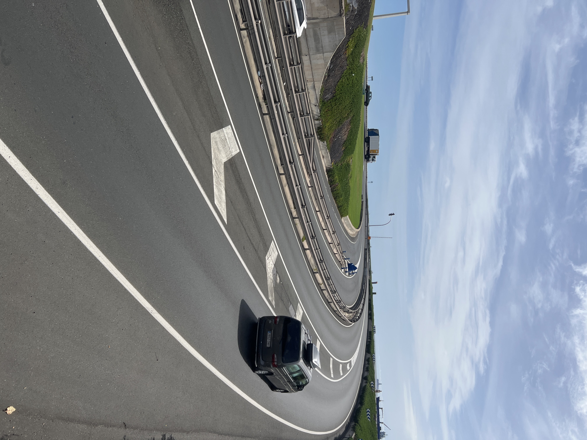 The image shows a section of highway in Las Palmas, Spain. A dark-colored vehicle is positioned on the left-hand side of the roadway, partially occupying the leftmost lane. The road has white lane markings and arrows indicating the direction of travel. A metal guardrail separates the highway from a grassy embankment. In the background, there are light-colored clouds against a blue sky. There are other vehicles on the road, including a truck and another car. The environment appears to be a bright, sunny day.