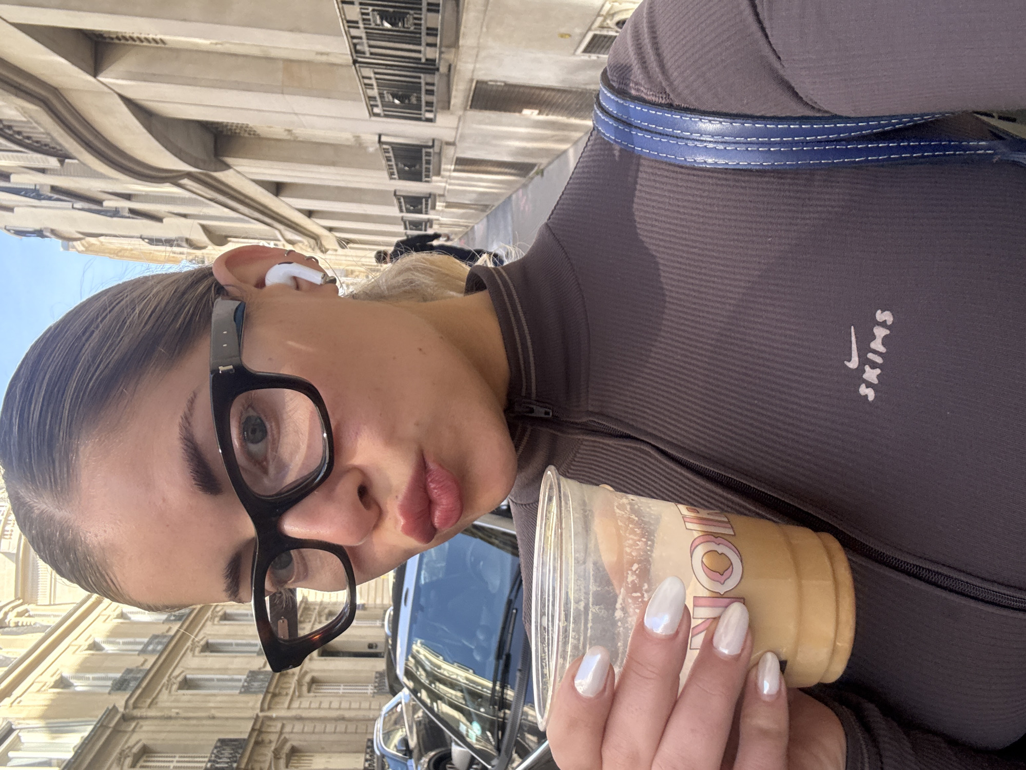 The photograph is a medium-range, vertical portrait of a woman with light skin, wearing black-rimmed glasses and white wireless earbuds in her left ear. She is holding a clear plastic cup containing a beige liquid, likely a coffee beverage, in her right hand. Her fingernails are painted with a pearlescent white polish.
She is wearing a dark brown, long-sleeved, ribbed top with a small white "SKIMS" logo on the right chest. A blue strap, possibly from a bag, is visible across her shoulder.
The background features Parisian architecture, including buildings with beige facades and numerous windows. A black car is partially visible behind the cup. The sky is a clear, pale blue, suggesting daylight. The location is likely Paris, France, given the architectural style.