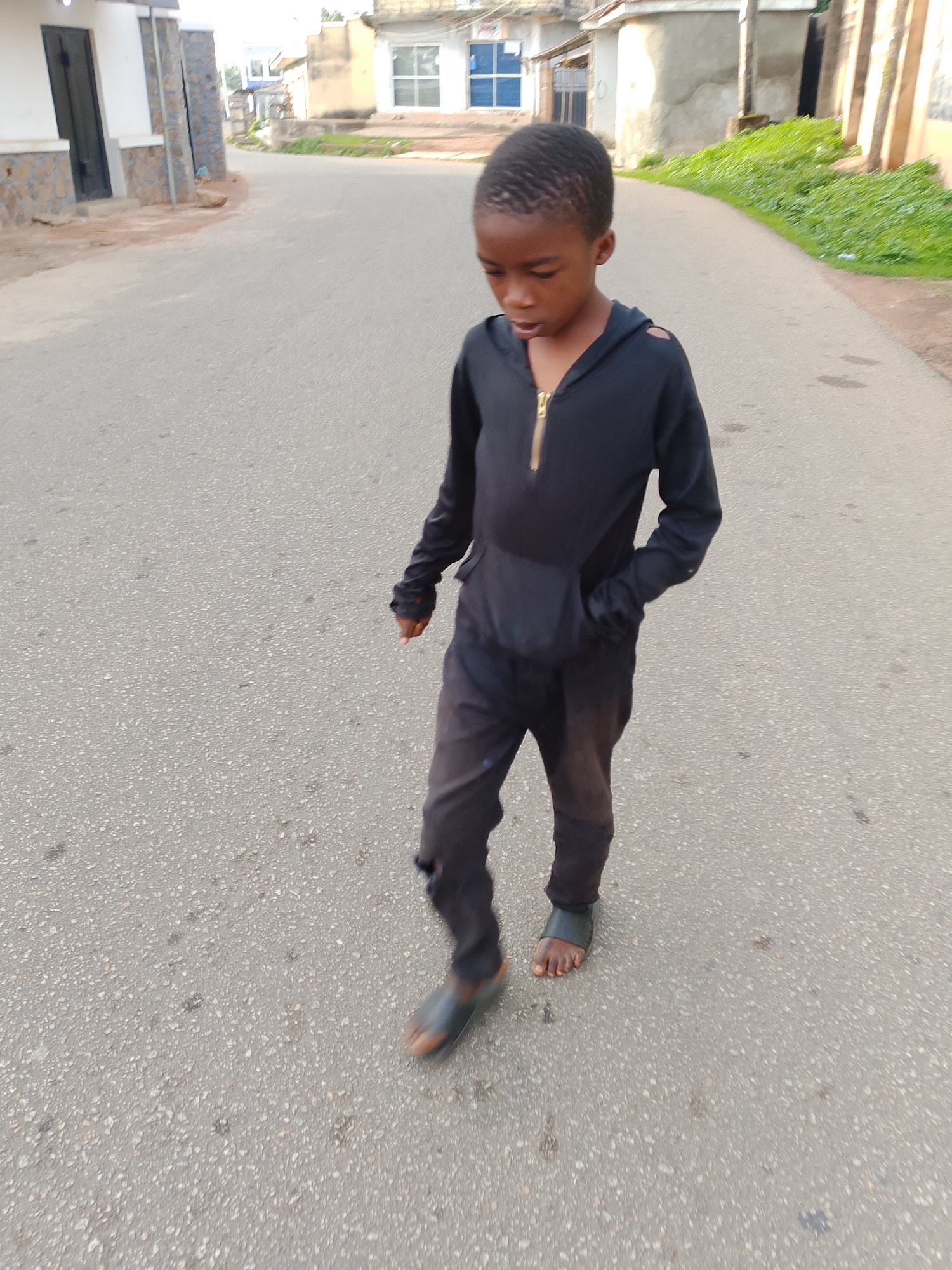 A young Black boy walks down a paved road in Jos, Nigeria. He is wearing a black hooded sweatshirt with a gold zipper and black pants. His right hand is in his pocket, and his left hand is partially visible. He is wearing dark sandals. The road is a medium gray color with some speckling. On the left side of the road, there is a building with white and stone-colored walls and a black door. Further down the road, there are other buildings, including one with a blue-windowed facade and another with rough, light-colored walls. Green foliage is visible along the right side of the road, behind the boy. The lighting suggests it is daytime, with some shadows present.