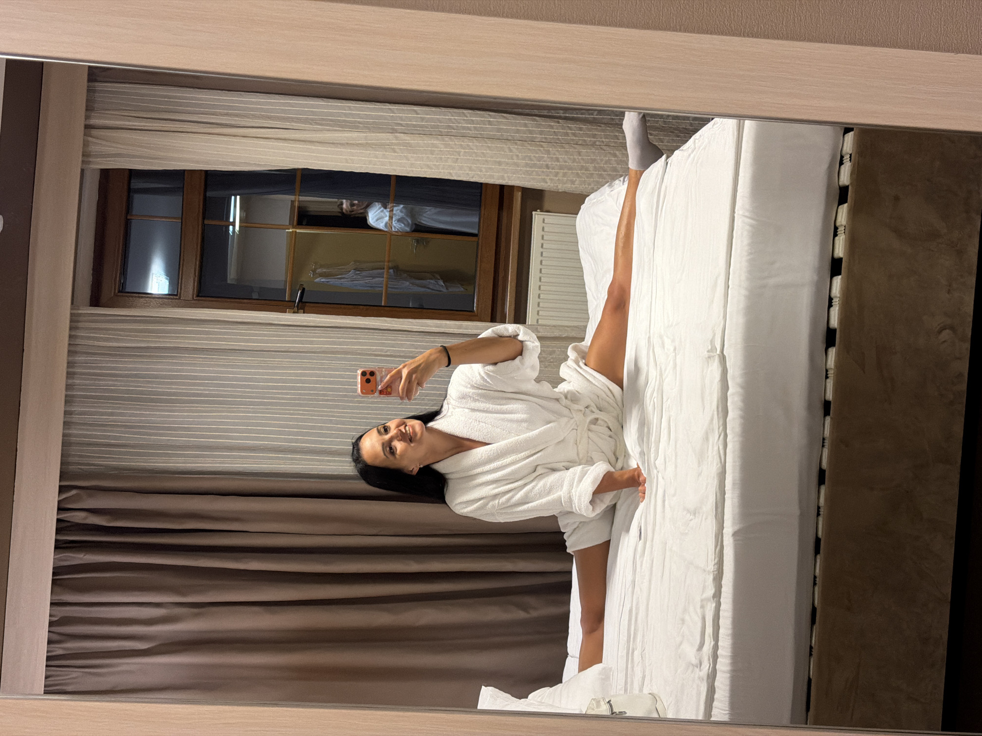 A woman in a white bathrobe is performing a split on a bed in a hotel room in Svetlogorsk, Russia. The room has brown curtains, a window with a reflection of another person, and a radiator. The woman is holding a smartphone and taking a selfie.