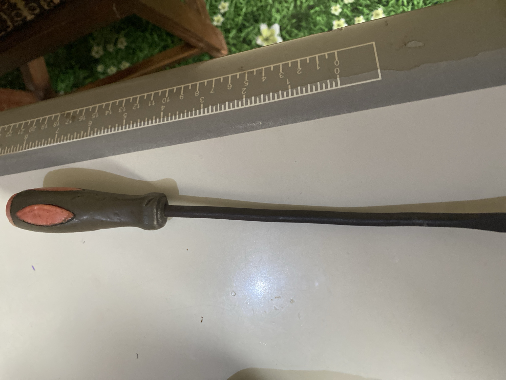 The image shows a close-up, overhead view of a pry bar with a dark handle featuring a red insert, lying on a flat, light-colored surface. To the right of the pry bar, a ruler with markings in centimeters and millimeters is visible, resting on a patterned surface with green and white floral motifs, suggesting it is placed on a rug or similar material. The ruler extends towards the right edge of the frame, where a dark wooden chair leg is partially visible, along with more of the patterned surface. The lighting suggests an indoor setting. There are no people present in the image, and no specific details are provided to indicate the location within Jauro Yinu, Nigeria.