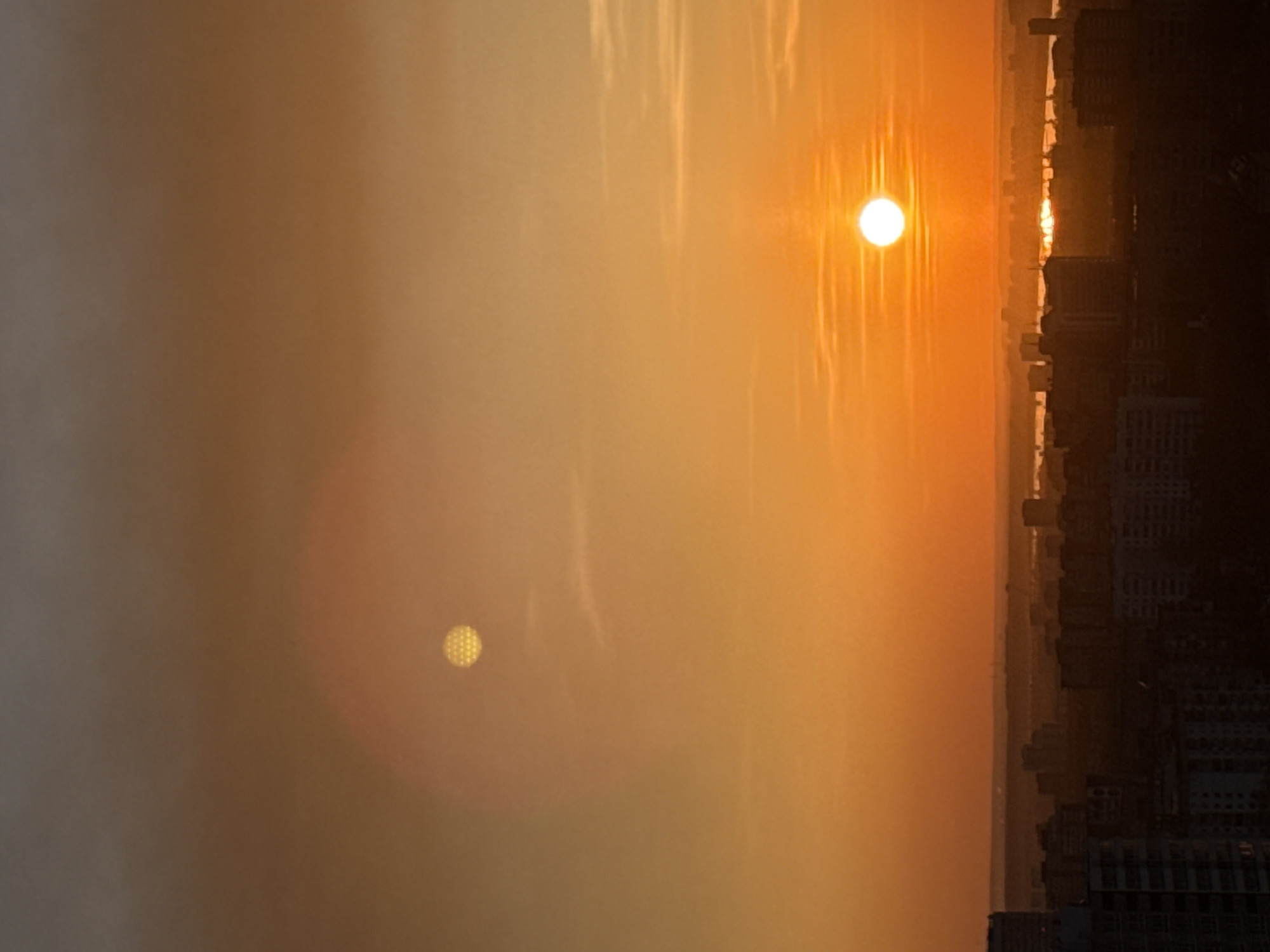 The image depicts a cityscape obscured by a thick, orange haze, likely due to smoke or pollution. The sun is visible as a bright orange orb with visible rays, and a fainter, blurry reflection is seen lower in the image. The silhouette of buildings is visible on the right side of the image, indicating a city setting. The image is oriented sideways. Due to the limited information provided, the exact location within Perm, Russia, cannot be determined.