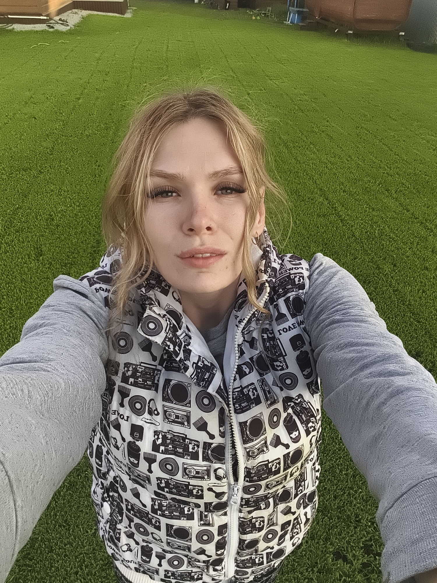 A young woman with blonde hair, styled casually around her face, is depicted in a high-angle selfie taken outdoors in Русский Вожой, Russia. She looks directly towards the camera with a neutral expression. She wears a light grey long-sleeved top underneath a white puffy vest. The vest features a repetitive black graphic pattern composed of retro musical equipment, including boomboxes, turntables, headphones, and vinyl records, along with some abstract shapes. A zipper is visible down the front of the vest.
The background is dominated by vibrant green artificial turf, suggesting a modern outdoor area like a backyard or recreation space. In the upper background, blurred structures, possibly parts of a building or fence, are faintly visible. The scene is brightly lit, indicating daytime conditions, possibly late afternoon or early evening, under clear skies. Some partial and reversed text, such as "TOAE" and "EWO" (or "MOB" reversed), is visible within the vest's pattern, but not fully decipherable as complete words.