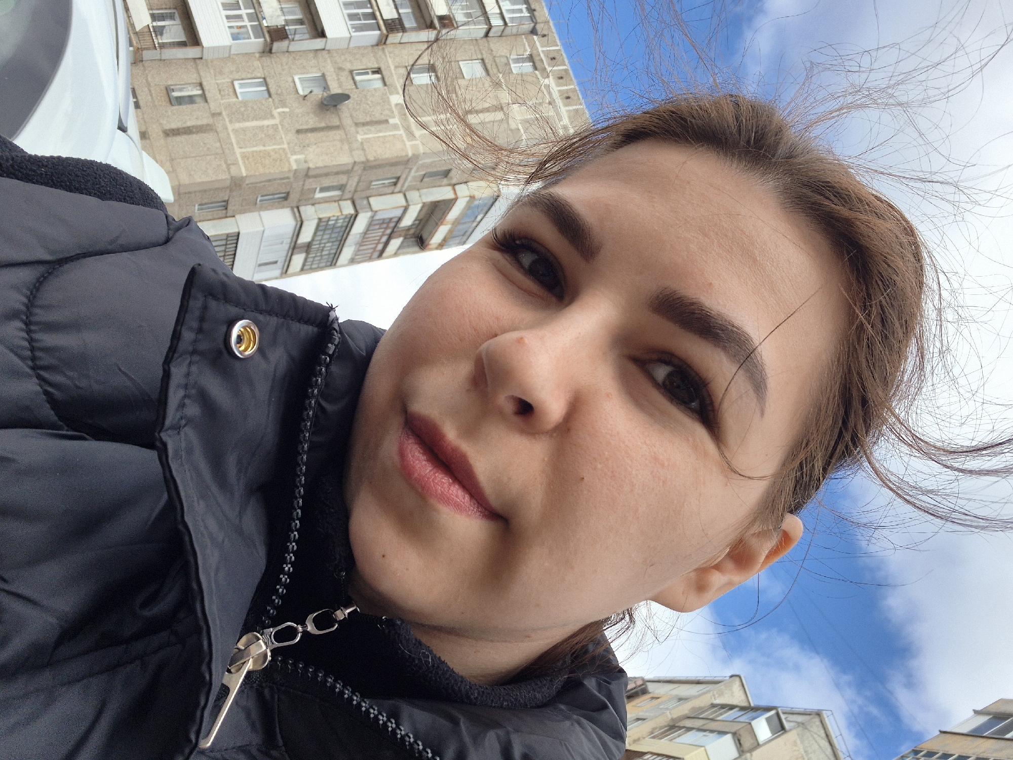 A young woman is taking a selfie outdoors, likely on a street in Tobolsk, Russia, as evidenced by the typical Soviet-era apartment buildings in the background. The woman is wearing a black puffy jacket, zipped up. Wisps of her brown hair blow in the wind. The sky is visible behind her.