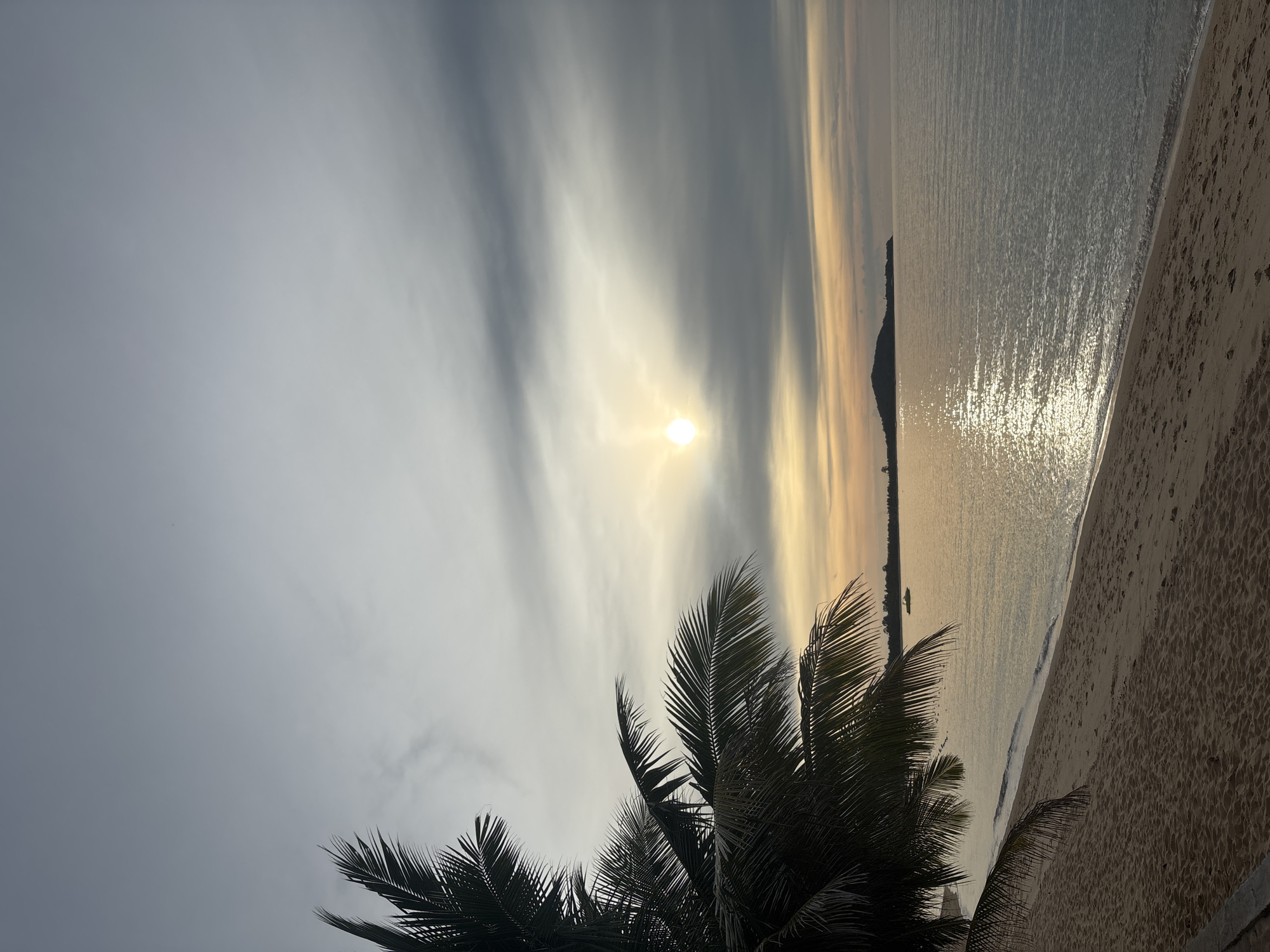 The image depicts a serene beach scene at dusk in Baan Bo Phut, Thailand. A prominent palm tree frond juts into the frame from the left, its dark silhouette contrasting with the muted colors of the sky. The sky is a gradient of grey and pale orange, with the sun visible as a bright, diffused orb peeking through a band of clouds. The ocean is calm, reflecting the sky's colors and the sunlight in shimmering streaks. The gentle waves lap onto a sandy beach, which shows some footprints. In the distance, a small, dark island or peninsula is visible on the horizon. A solitary boat is anchored on the water. The overall atmosphere is tranquil and still, suggesting the end of the day.