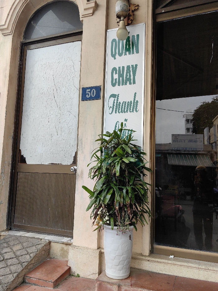 The scene depicts the entrance of a building, likely a vegetarian restaurant, located at 50, Thuận Hóa Ward, Vietnam. The building facade is a faded light beige. To the left, a brown-framed door features a lower panel with vertical ridging and an upper glass panel obscured by a white, cracked, opaque film; a section of this film is missing from the bottom left, revealing a dark interior. Above the door, a small, dark, semicircular window is visible within an arched alcove. A silver-colored doorknob is centered on the door. To the right of the door, a blue rectangular sign with the white number "50" is affixed to the wall. Above this, an ornate, distressed wall-mounted light fixture with a single clear-glass bulb points downwards. Below the light fixture, a tall, vertical white sign with green Vietnamese text, primarily "QUAN CHAY Thanh" (Vegetarian Restaurant Thanh), is mounted. A large, dark green leafy plant, with some withered foliage and small white and yellow blossoms, stands in a white, textured cylindrical ceramic planter positioned on the second of two reddish-brown tiled steps. These steps lead up to the entrance, with a diagonally checkered reddish-brown tiled ramp visible to the far left. To the right of the plant, a large, dark-tinted window reflects the street scene opposite, including a building with a green awning and faint Vietnamese text, and the clear reflection of a person wearing a light-colored helmet, holding a device, presumably the photographer. No direct interactions or distinct actions are visible within the frame, beyond the implied presence of the photographer.