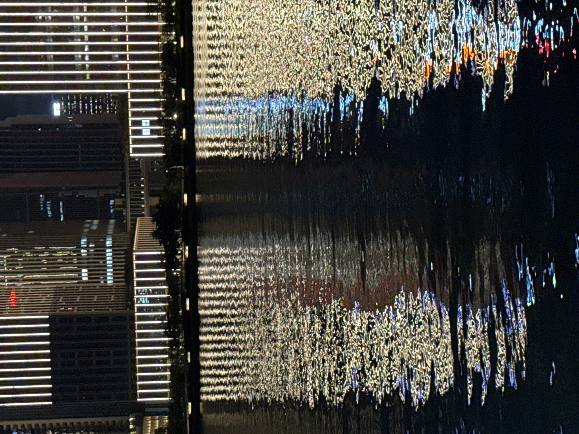This is a nighttime photograph depicting an urban waterfront scene in Tianhe District, China. Multiple high-rise buildings are visible in the background, characterized by prominent vertical bright yellow-white light strips on their facades. Some buildings exhibit minor red and blue illuminated elements. A dark strip of land, likely a riverbank, separates the structures from a body of water in the foreground, featuring silhouetted vegetation and smaller, horizontal light sources. The water's surface displays fragmented and elongated reflections of the building lights, caused by ripples. These reflections are predominantly bright yellow-white, with discernible streaks of blue, red, and orange light scattered within the patterns. No discernible individuals are present in the image.