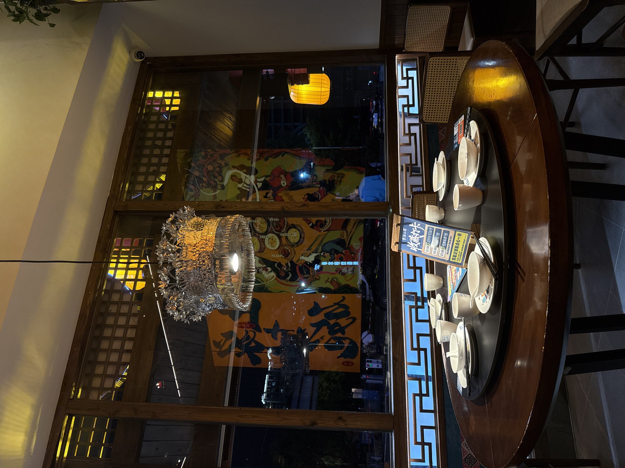 The image captures an indoor setting in Zhangjiajie, China, likely a restaurant. A round, dark wood table is set with white ceramic tea cups and saucers, and chopsticks, suggesting a tea service. A sign in Chinese is positioned at the table. The backdrop features a large window showcasing decorative Chinese artwork and city lights. A modern, metallic chandelier hangs above the table. The overall lighting is a mix of natural light from the window and artificial illumination from the chandelier and a distant yellow lantern visible outside. The environment appears upscale with intricate woodwork and attention to detail.