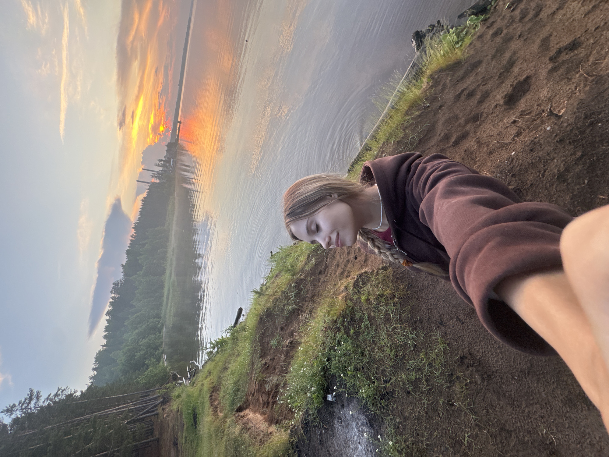 A young woman with blonde hair in a braid, wearing a maroon hoodie, is taking a selfie on a sandy bank overlooking a calm body of water. The sun is setting, casting a warm orange glow across the sky and reflecting on the water. A dense forest lines the opposite bank, with industrial structures visible in the distance. The foreground shows the sandy terrain with scattered grass and small white flowers. The image is captured from a high angle, looking down at the woman. The overall scene suggests a serene, natural landscape. This appears to be a location along the Kama River in or near the city of Chaikovsky, Russia, known for its industrial sites and surrounding natural beauty.