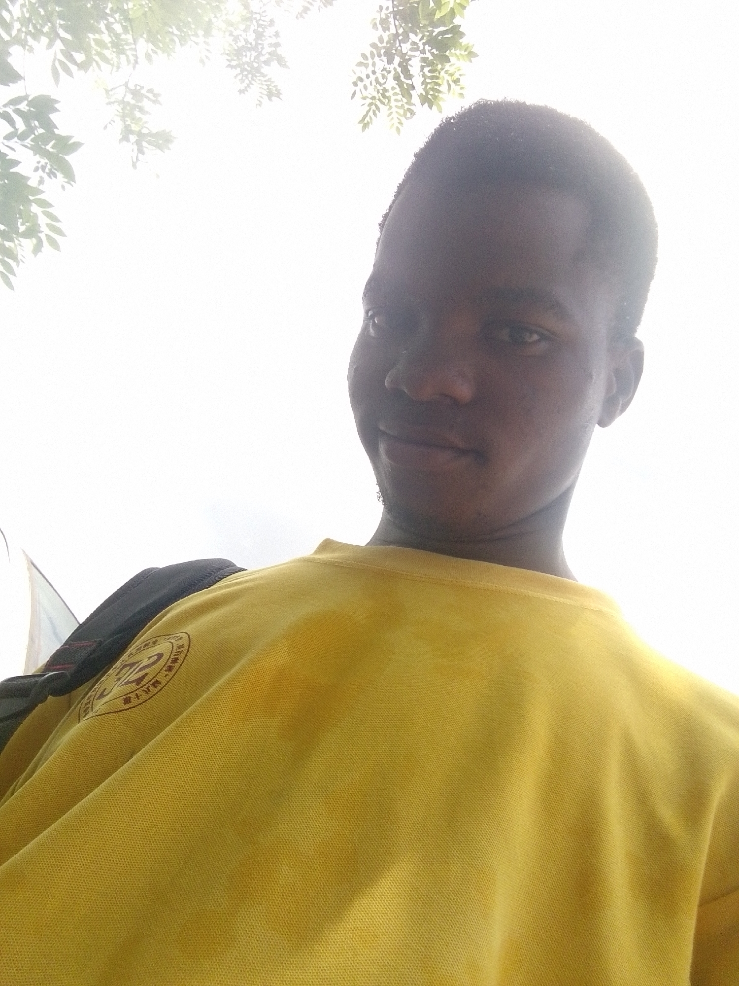 A medium-shot, low-angle, underexposed, outdoor image shows a person wearing a yellow shirt with a patch on the left breast and a black backpack on their left shoulder. The person has dark skin, short hair, and is looking at the camera with a slight smile. The background is a bright, overcast sky with some green leaves visible at the top of the frame. The location is Eleweran, Nigeria.