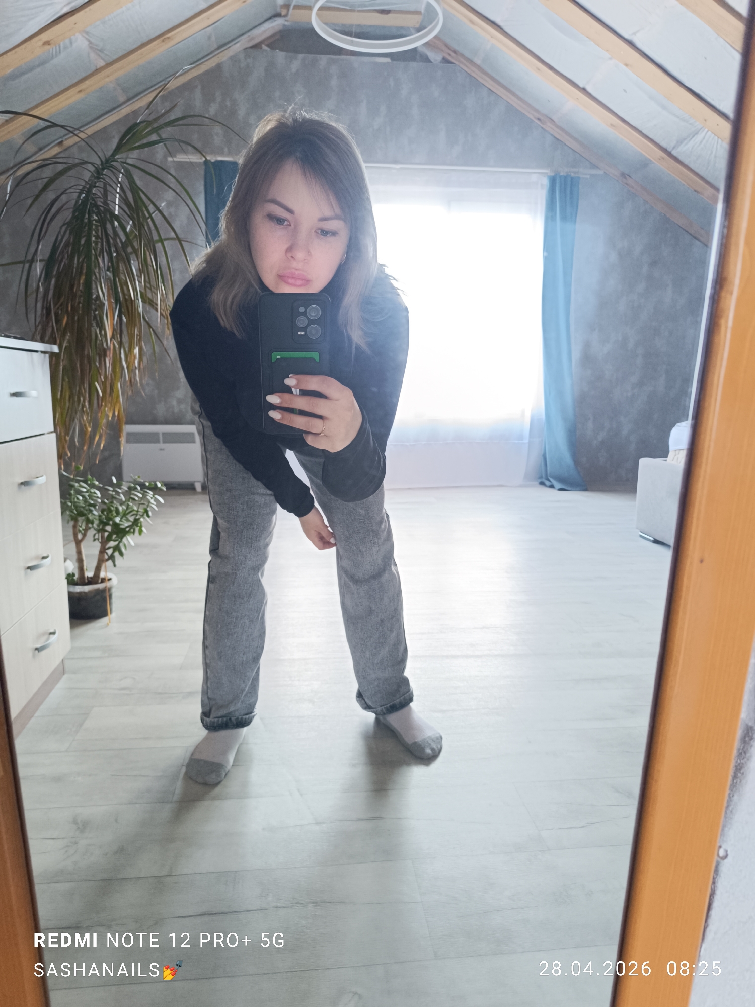 A woman is captured in a mirror selfie inside a room, likely an attic or loft space, in Chaikovsky, Russia. She has light brown hair, wears a black long-sleeved top, light grey jeans, and grey socks, and displays a slight pout on her face. She holds a black smartphone, with a green card visible in its case, using it to take her reflection in the wooden-framed full-length mirror.
The room features a sloped ceiling with exposed wooden beams and white insulation. The walls are a textured grey, and the floor is a light wood or laminate. To the left, a tall, leafy houseplant and a smaller jade plant sit near a light-colored dresser with silver handles. A white electric radiator is visible below a bright window adorned with light blue curtains, suggesting daylight. A modern, circular light fixture hangs from the ceiling, and a light-colored armchair is partially visible in the background.
The timestamp "28.04.2026 08:25" indicates the photo was taken in the morning. Text visible at the bottom left corner reads "REDMI NOTE 12 PRO+ 5G" and "SASHANAILS💅".