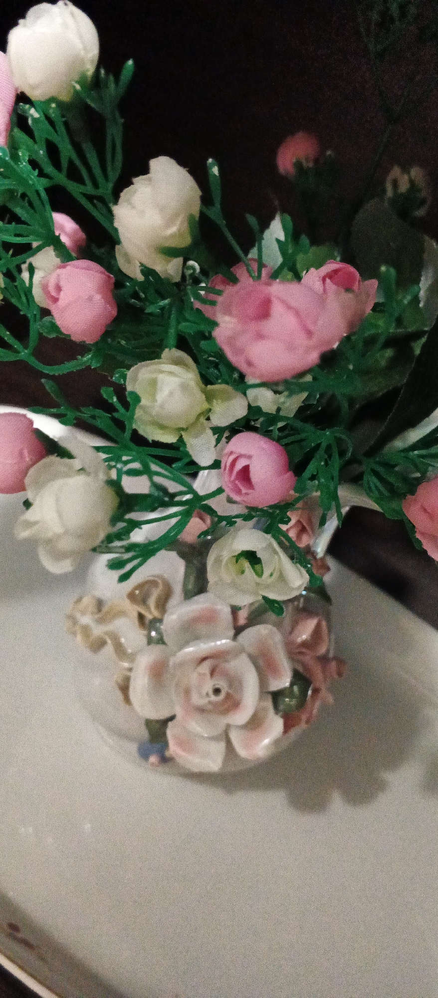 A close-up, low-angle, vertical shot shows a decorative vase filled with artificial flowers. The vase appears to be ceramic and is white with pink and beige floral embellishments. The flowers are a mix of pink and white, with green plastic leaves and stems. Some of the flowers are tightly budded, while others are more open. The background is dark and out of focus, making it difficult to discern any specific details of the environment. The lighting is dim, casting shadows and creating a soft, slightly grainy image. There are no people or animals visible in the image. It is not possible to determine the location in Berehove, Ukraine, from the visual information provided.